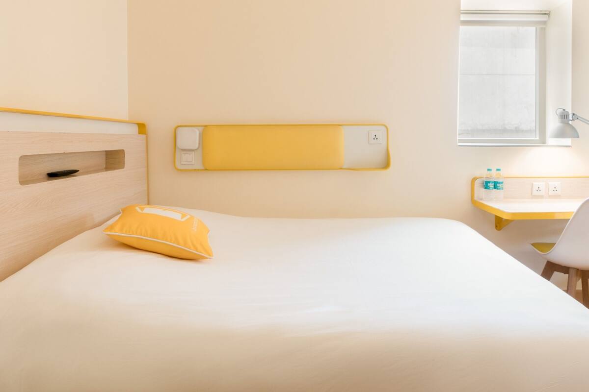 Een hotelkamer met een bed, nachtkastje met lamp, klein bureau en raam met uitzicht.