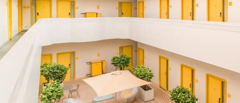 Un vestíbulo de hotel de varios niveles con puertas amarillas, un área de descanso central con una mesa y sillas, y plantas en macetas.