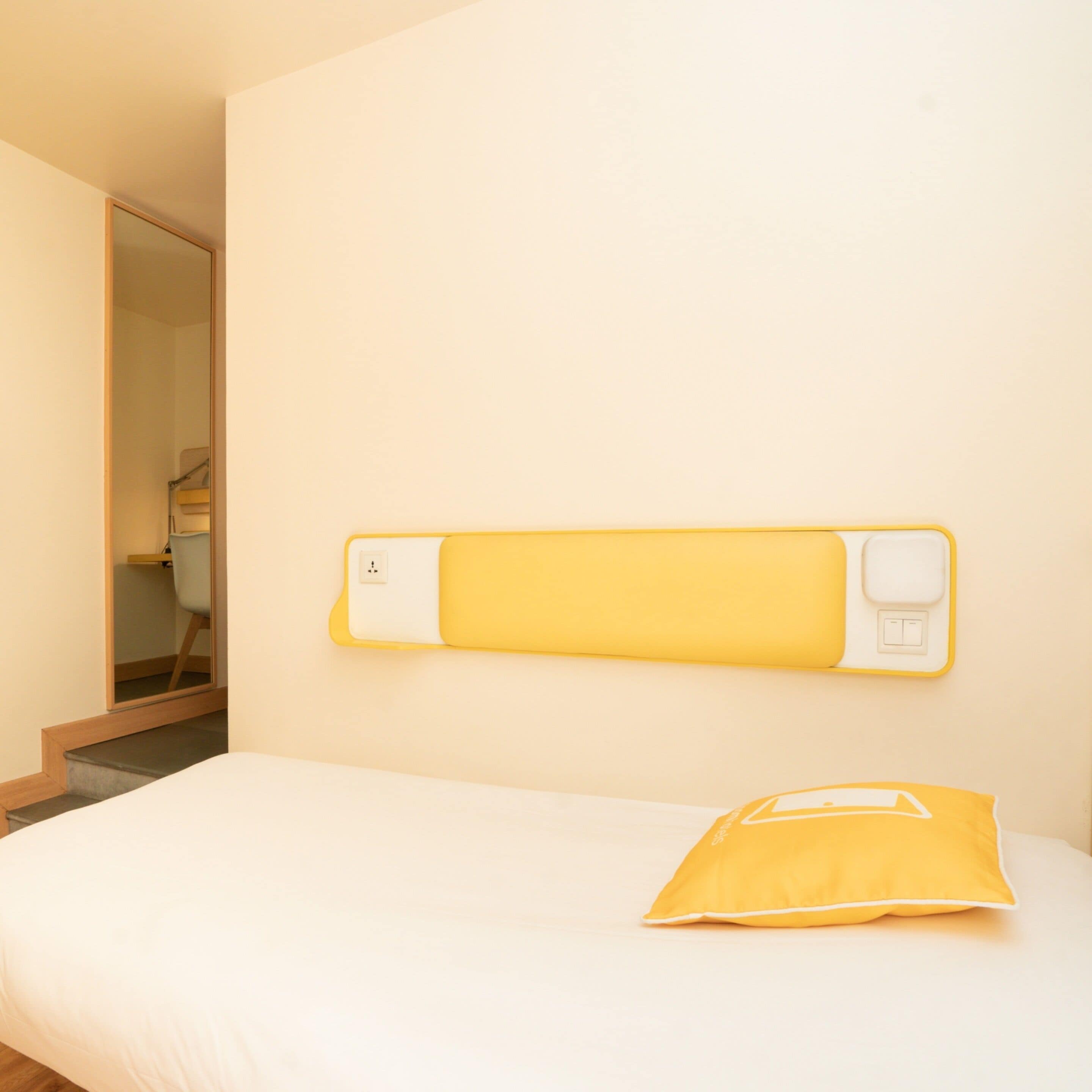A bedroom with a bed, a yellow pillow, and a wall-mounted power outlet.