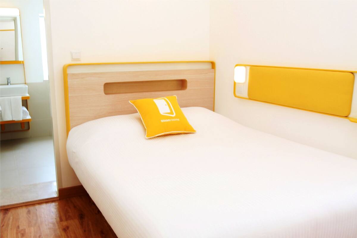 Een hotelkamer met een houten hoofdeinde, een wit bed en een geel kussen met het logo 'Bloom Rooms'.