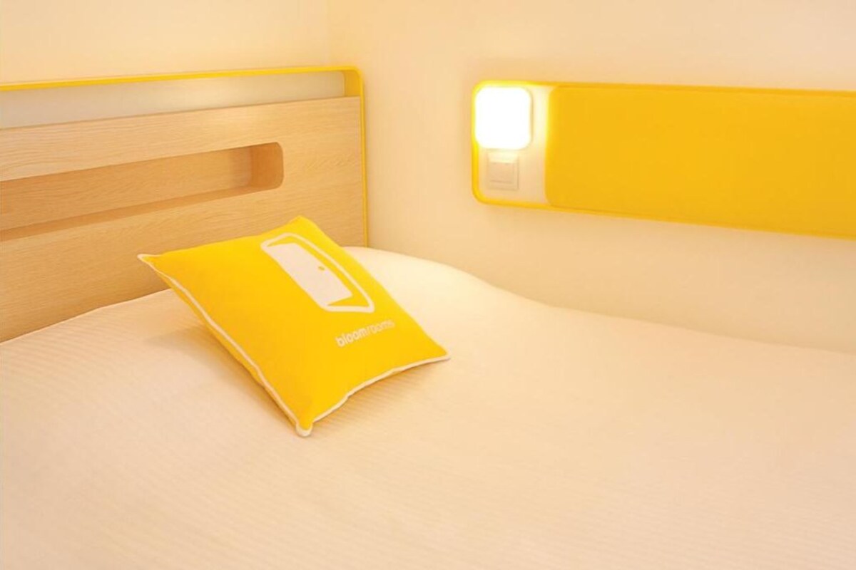 Un lit avec une tête de lit en bois, un oreiller jaune orné d’un logo blanc et une table de chevet avec une lampe.