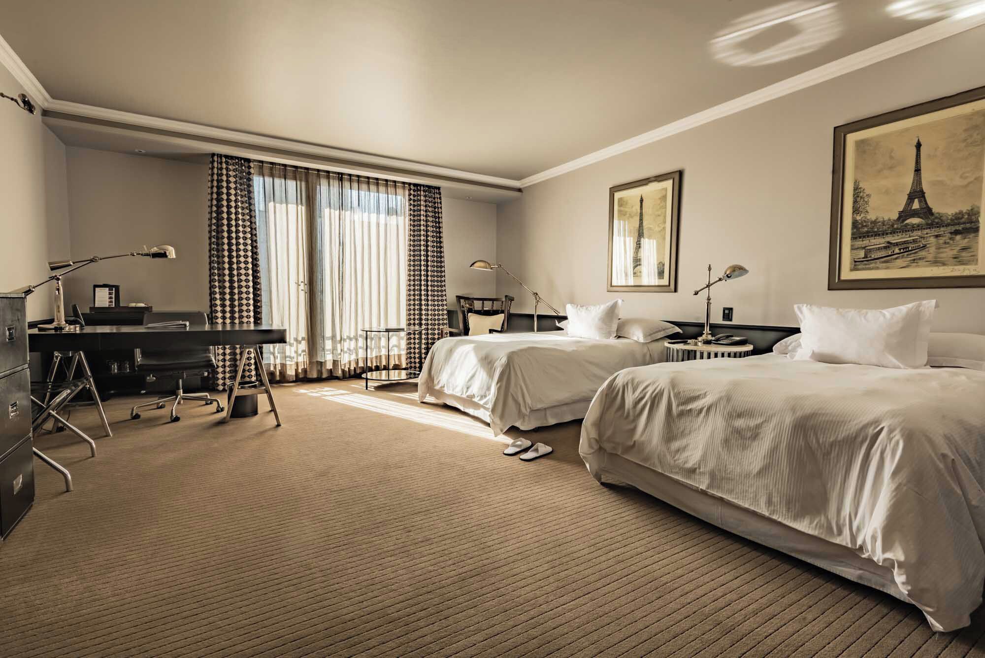 A hotel room with two beds, a desk, a chair, a lamp, and a large window with curtains.