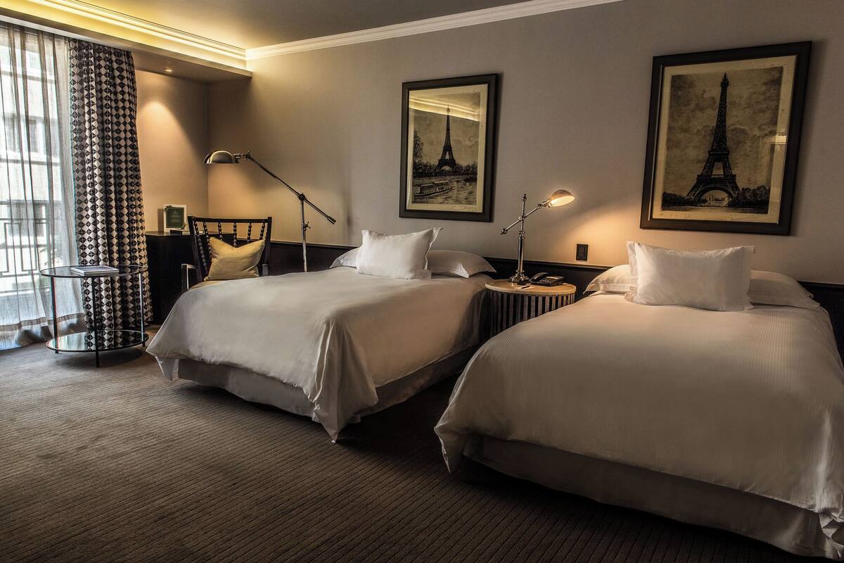 A hotel room with two beds, a desk, a chair, a lamp, and two framed pictures of the Eiffel Tower.