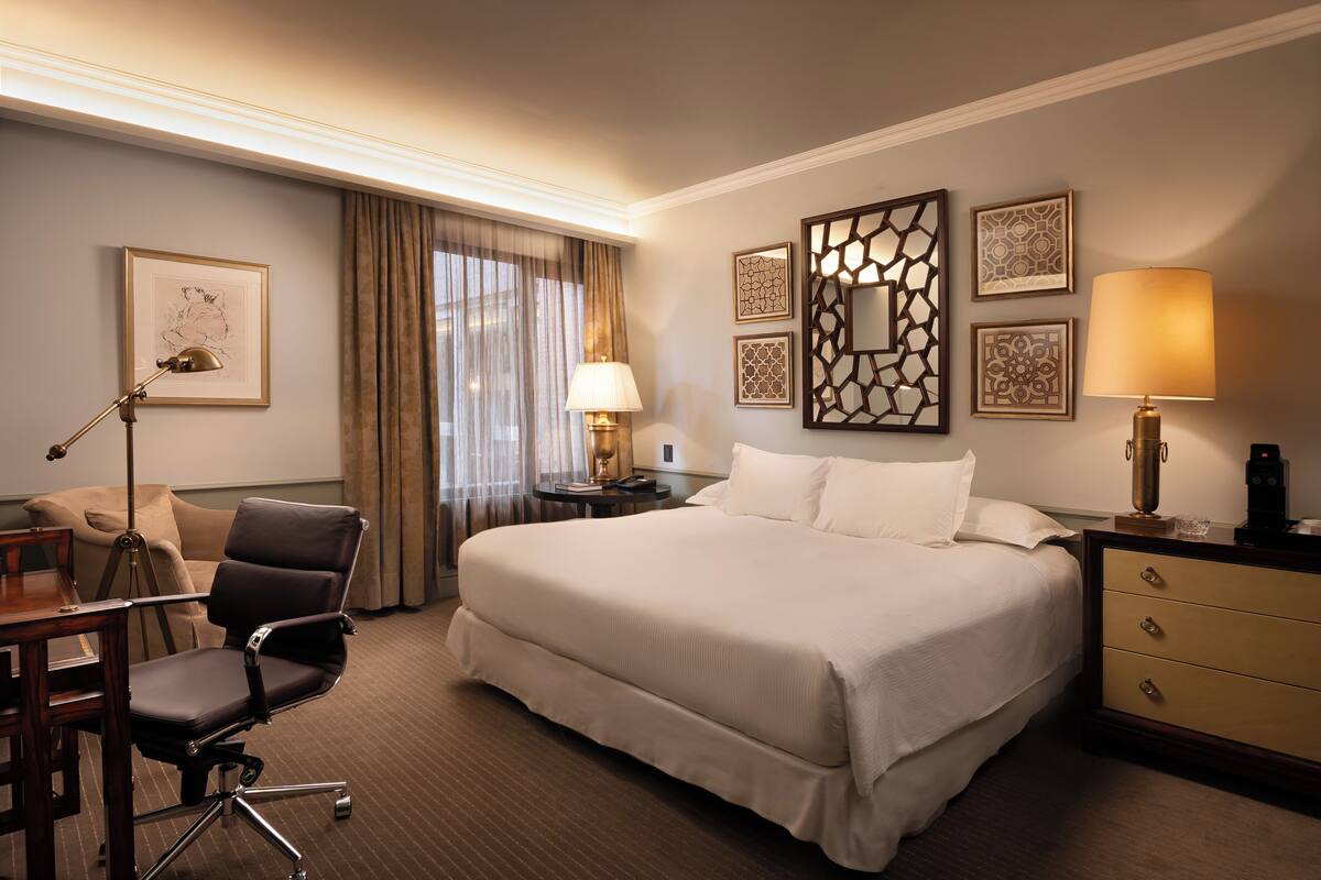 A hotel room with a large bed, a desk with a chair, a lamp, and framed artwork on the walls.