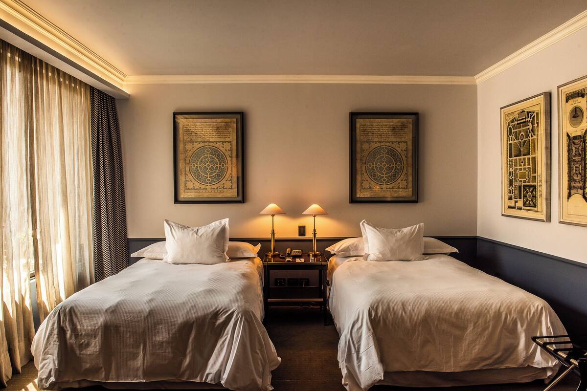 A hotel room with two single beds, a nightstand with lamps, and framed artwork on the walls.