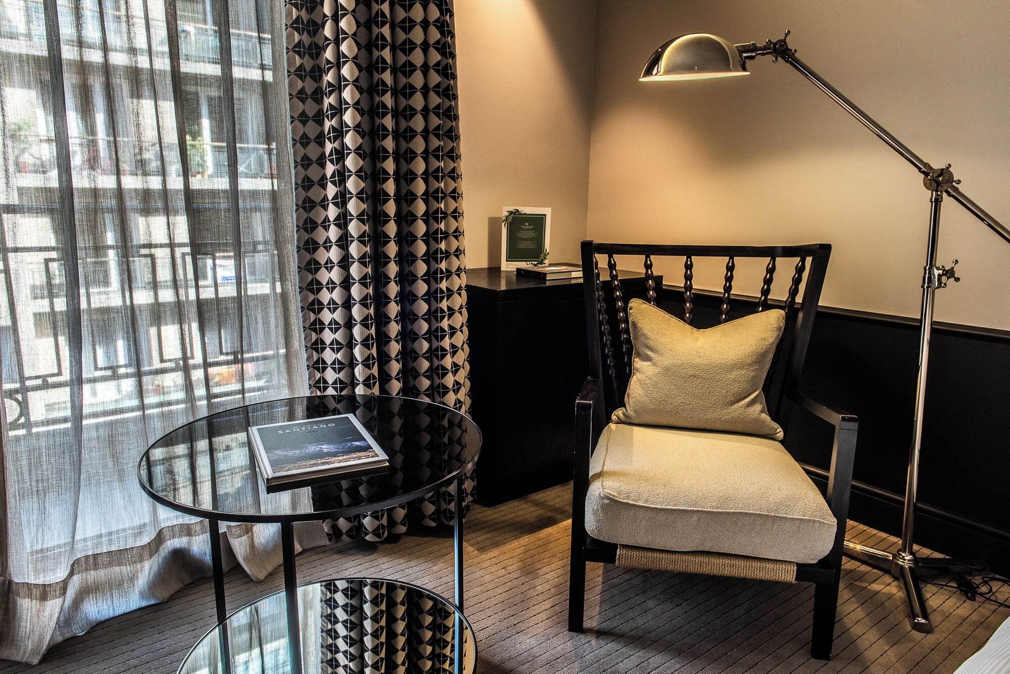 A room with a black and white patterned chair, a glass table with a book, a lamp, and a view through sheer curtains.