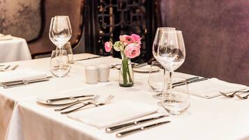 A dining table set with white tablecloth, silverware, and clear wine glasses. A vase with pink flowers is placed in the center.