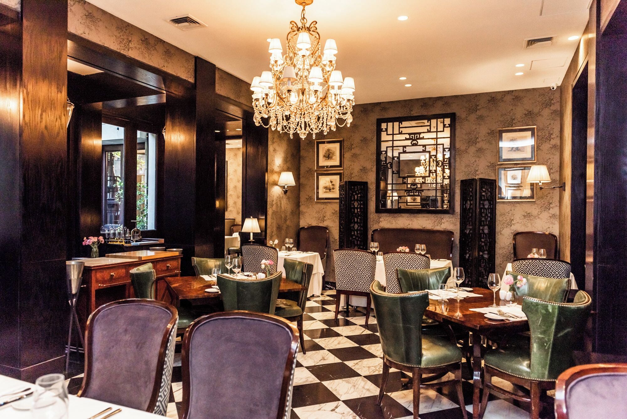 A restaurant interior with a checkered floor, patterned chairs, a chandelier, and framed artwork on the walls.