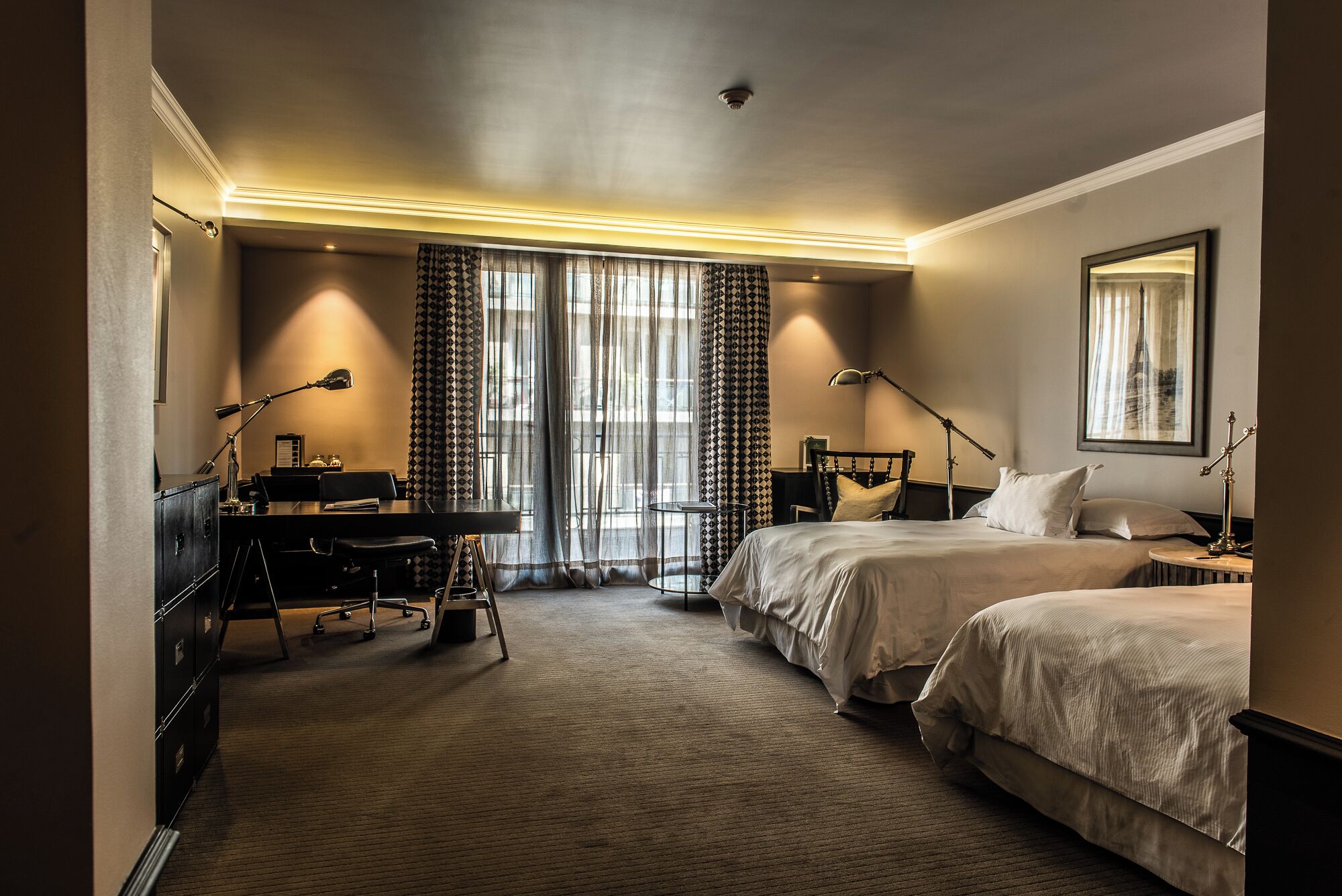 Habitación de hotel con dos camas, un escritorio con silla, una lámpara y un ventanal con cortinas.