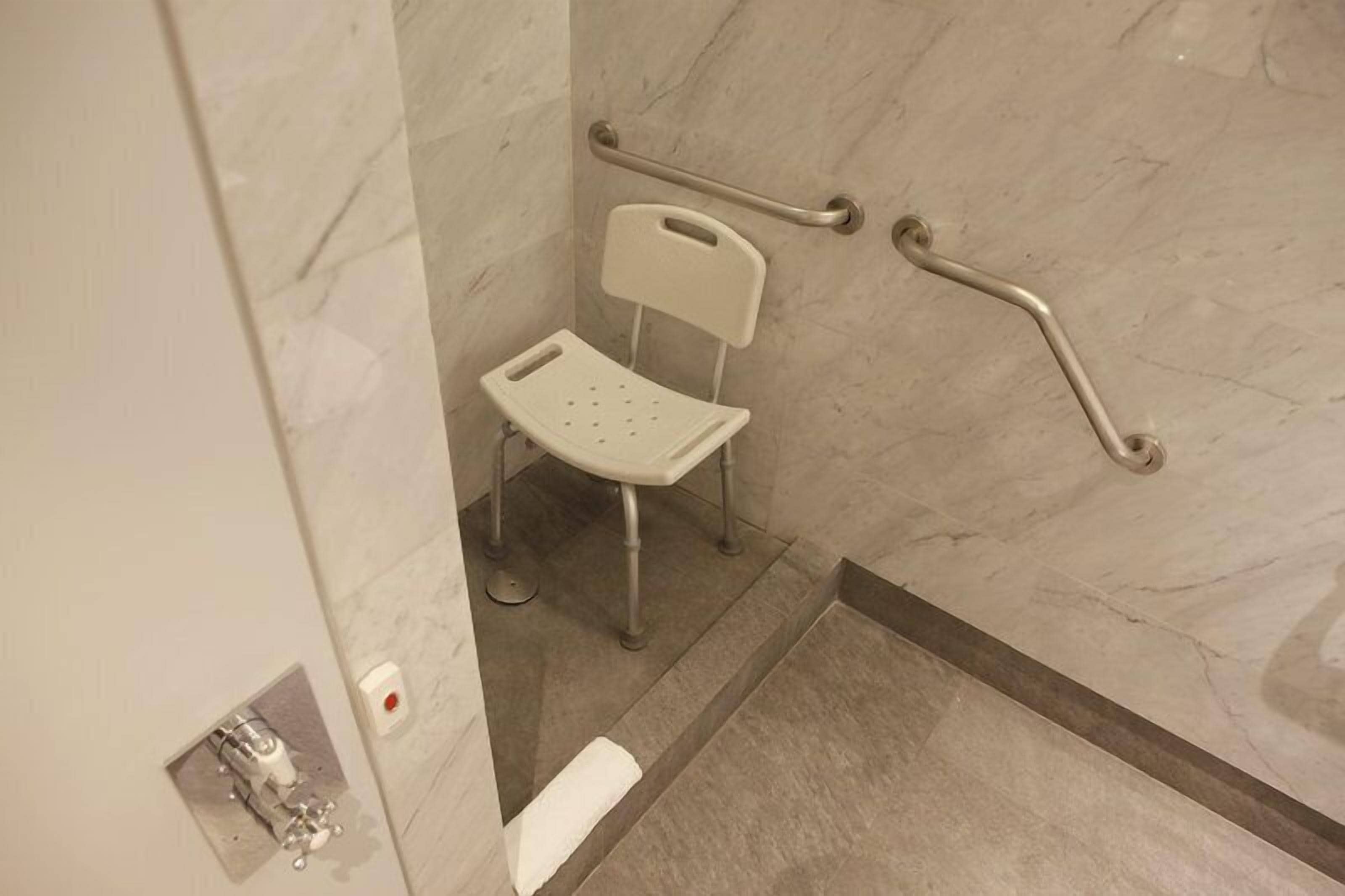 A bathroom with a white chair, grab bars, and a marble floor.
