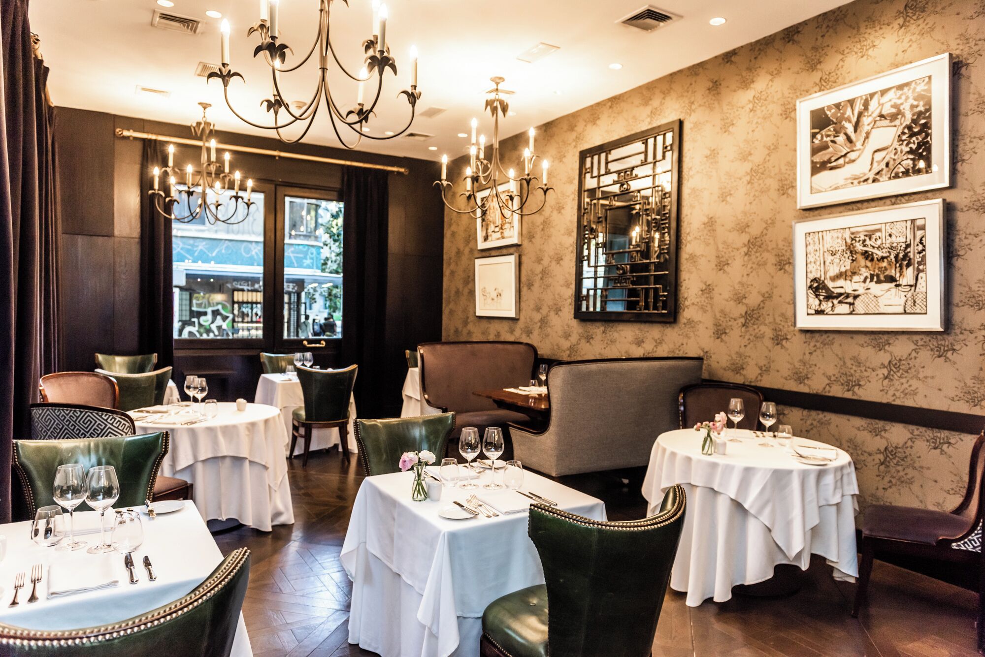 A restaurant interior with round tables, green upholstered chairs, chandeliers, and framed artwork on the walls.