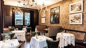 A restaurant interior with round tables, green upholstered chairs, chandeliers, and framed artwork on the walls.
