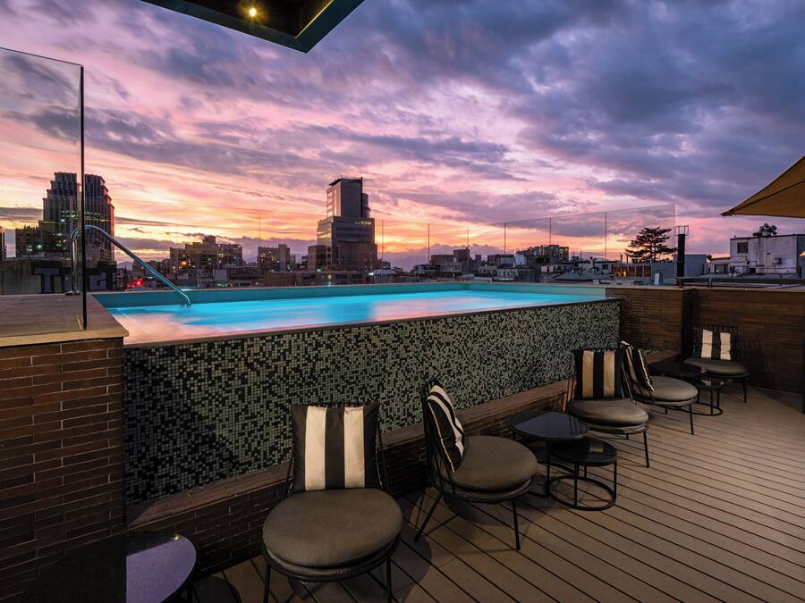 Un espace piscine sur le toit, offrant une vue sur les toits de la ville au coucher du soleil, avec un mur recouvert de carreaux, des sièges et une terrasse en bois.