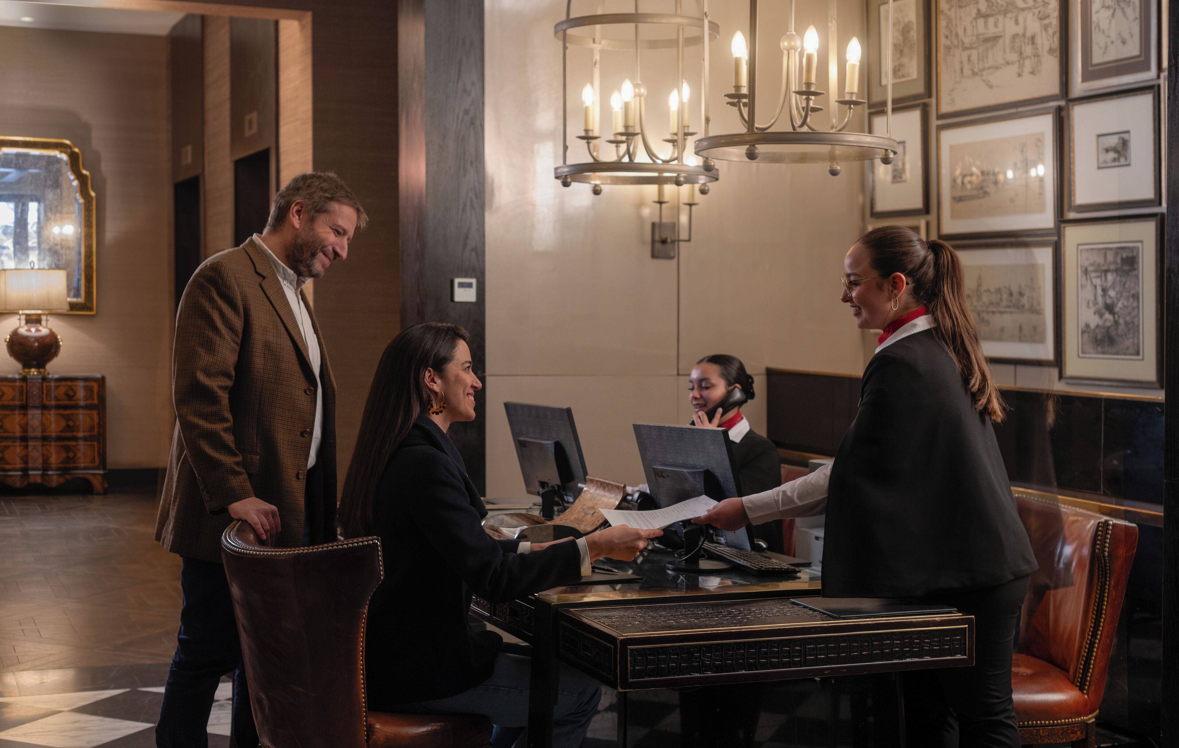 A lobby with a reception desk, chandeliers, framed artwork, and a check-in counter.