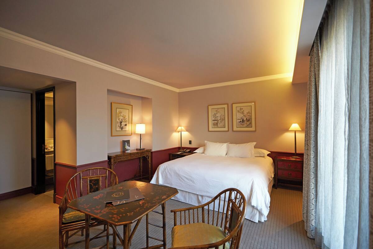 A hotel room with a bed, bedside tables, a desk with a chair, and a dining table.
