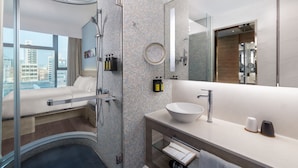 A modern bathroom with a glass-enclosed shower, a large mirror, a white sink, and a view of the city. - Lodgewood by Nina Hospitality丨Mong Kok (Kowloon)