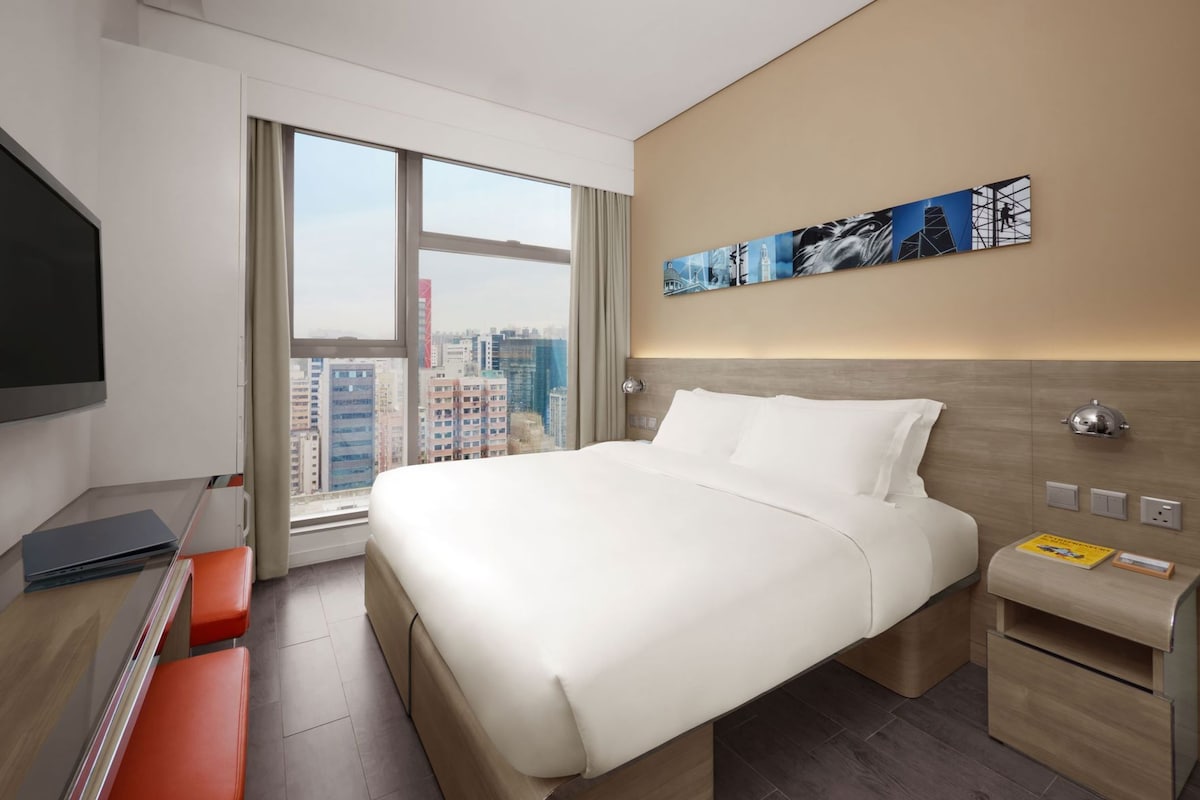 Ein Hotelzimmer mit einem großen Bett, einem Schreibtisch mit Laptop, einem Fernseher und Blick auf die Stadt.