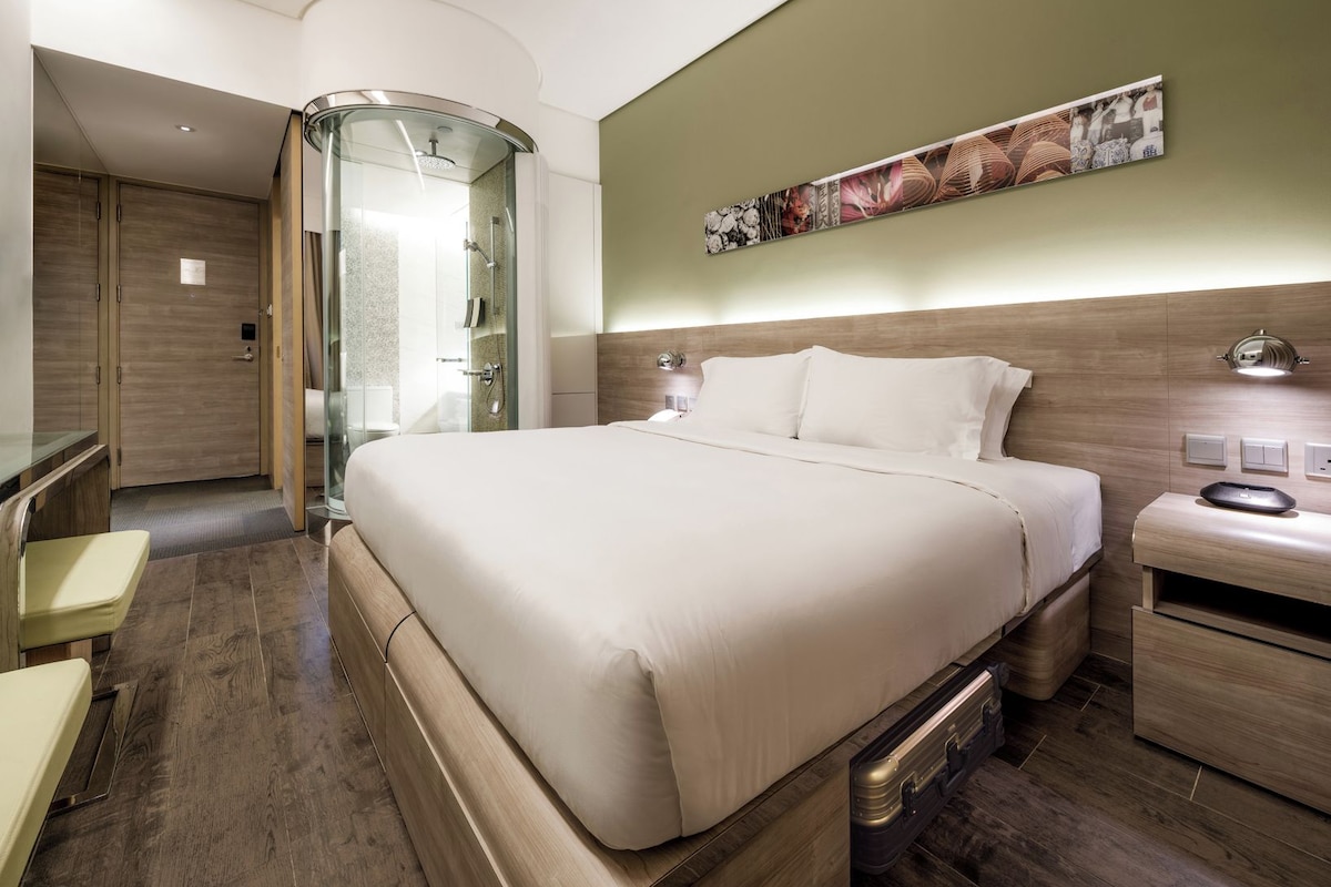 Ein Hotelzimmer mit einem großen Bett, einer gläsernen Dusche, einem Nachttisch und einem an der Wand hängenden Kunstwerk.