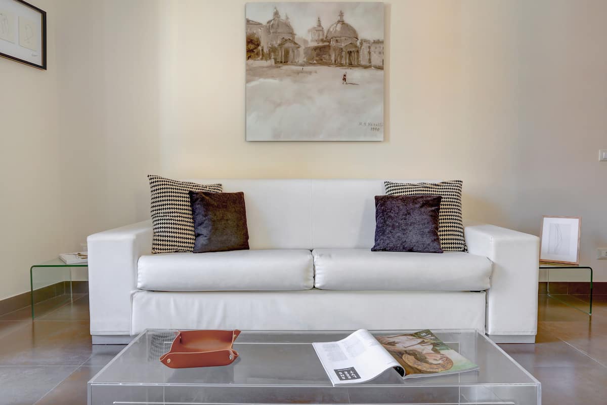A living room with a white sofa, two patterned throw pillows, a glass coffee table with a magazine and a brown object, and a painting of a historical building on the wall.