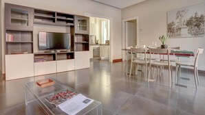 A modern living room with a flat-screen TV, built-in shelves, a dining area with chairs, and a kitchen in the background. - Colosseo Gardens - My Extra Home (Rome)