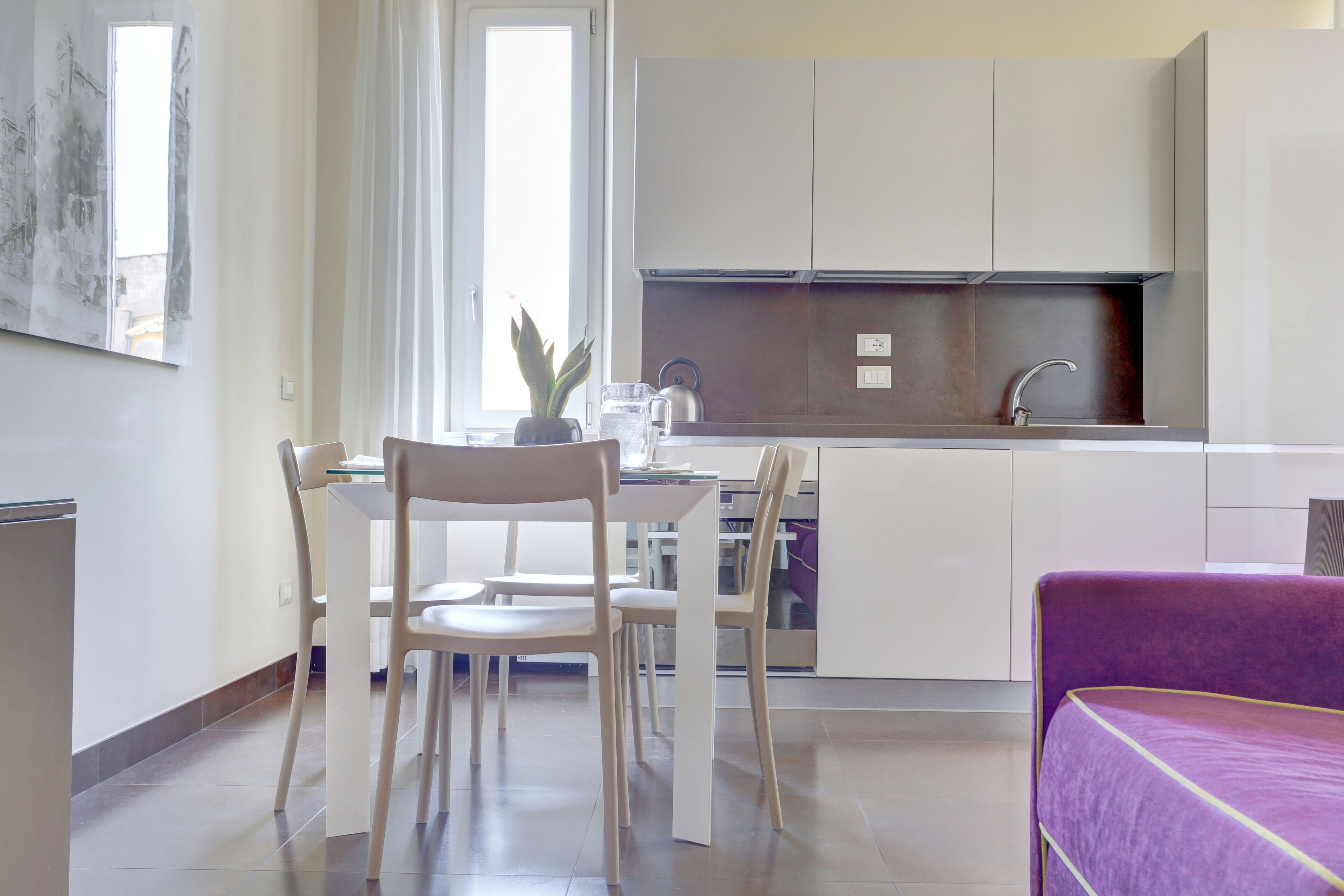 A modern kitchen with white cabinets, a built-in oven, and a sink. There is a dining area with a table and chairs, and a purple sofa in the foreground.