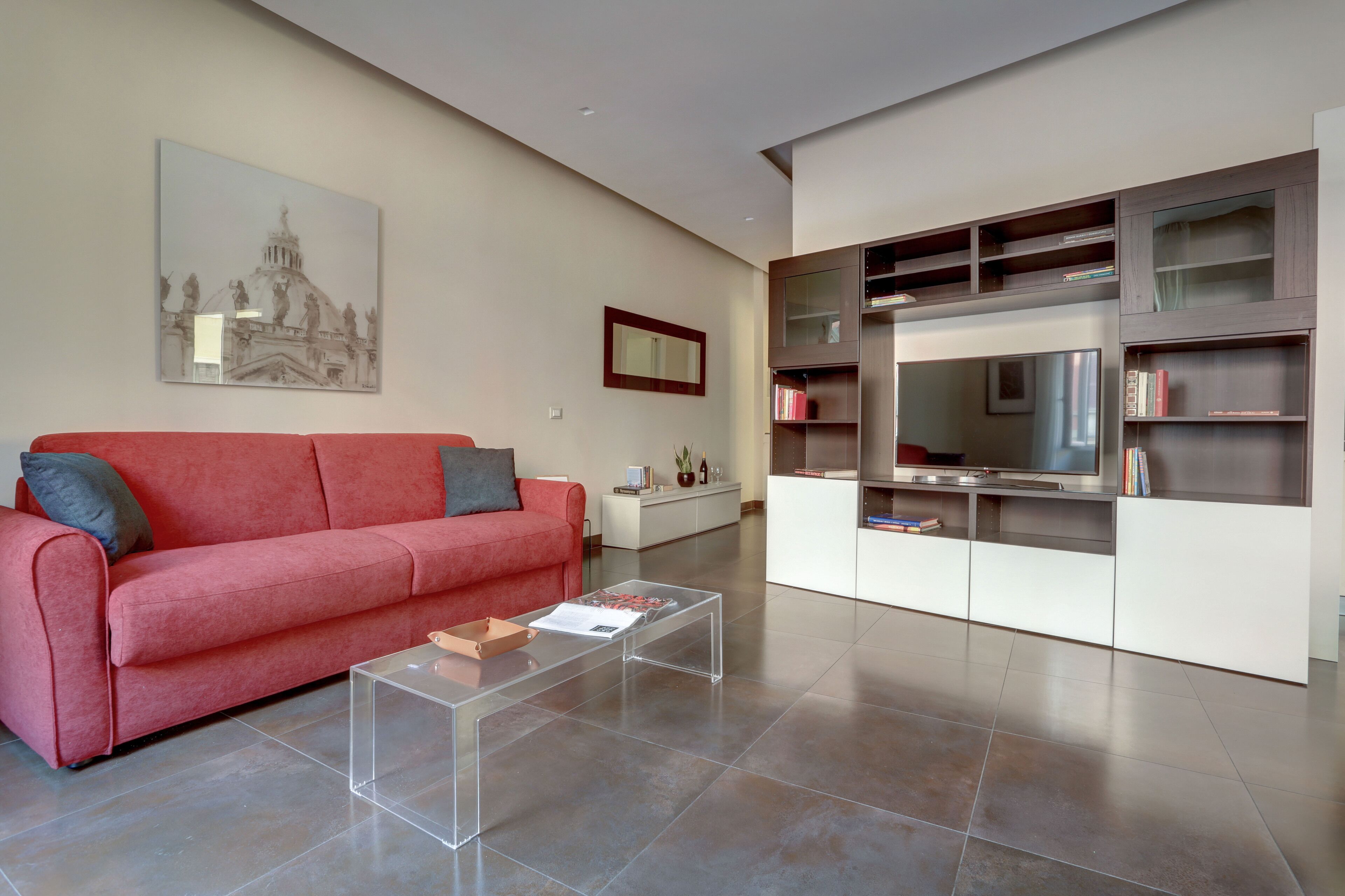 A modern living room with a red sofa, a clear coffee table, a large flat-screen TV, and built-in shelving with books.