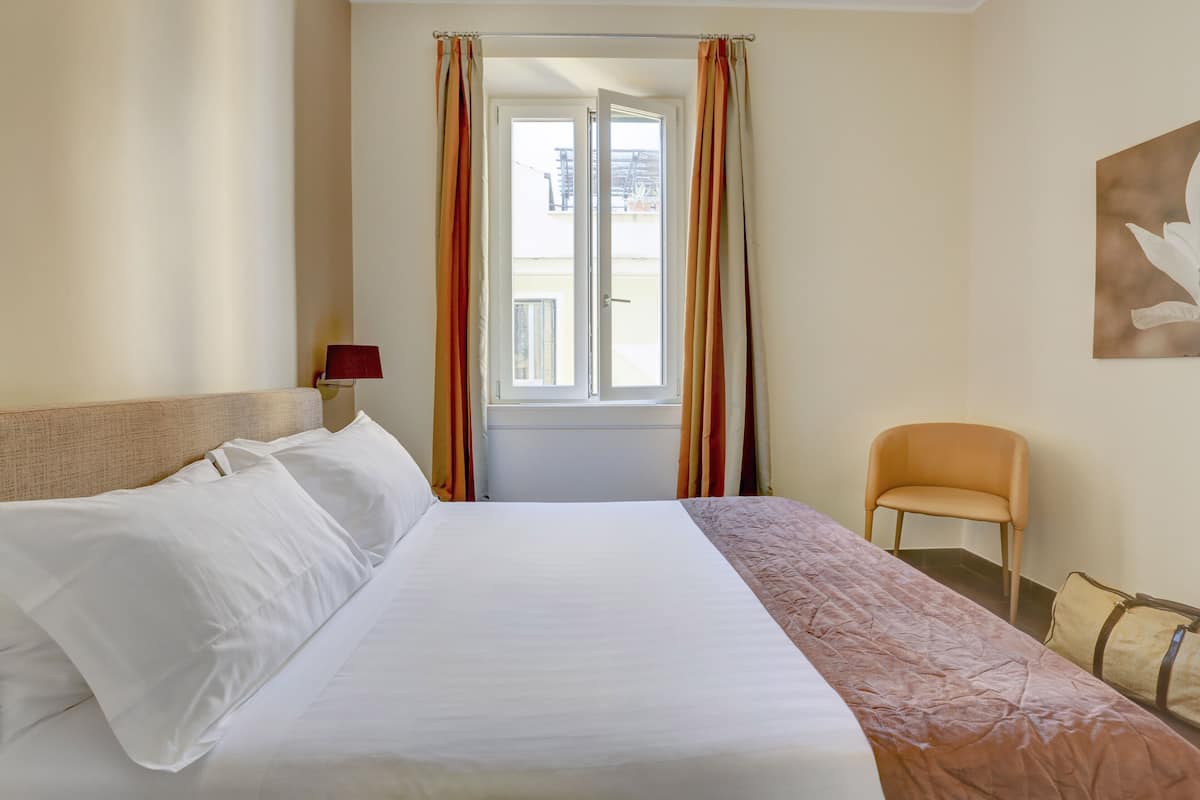 A hotel room with a large bed, a chair, a window with curtains, and a wall-mounted lamp.