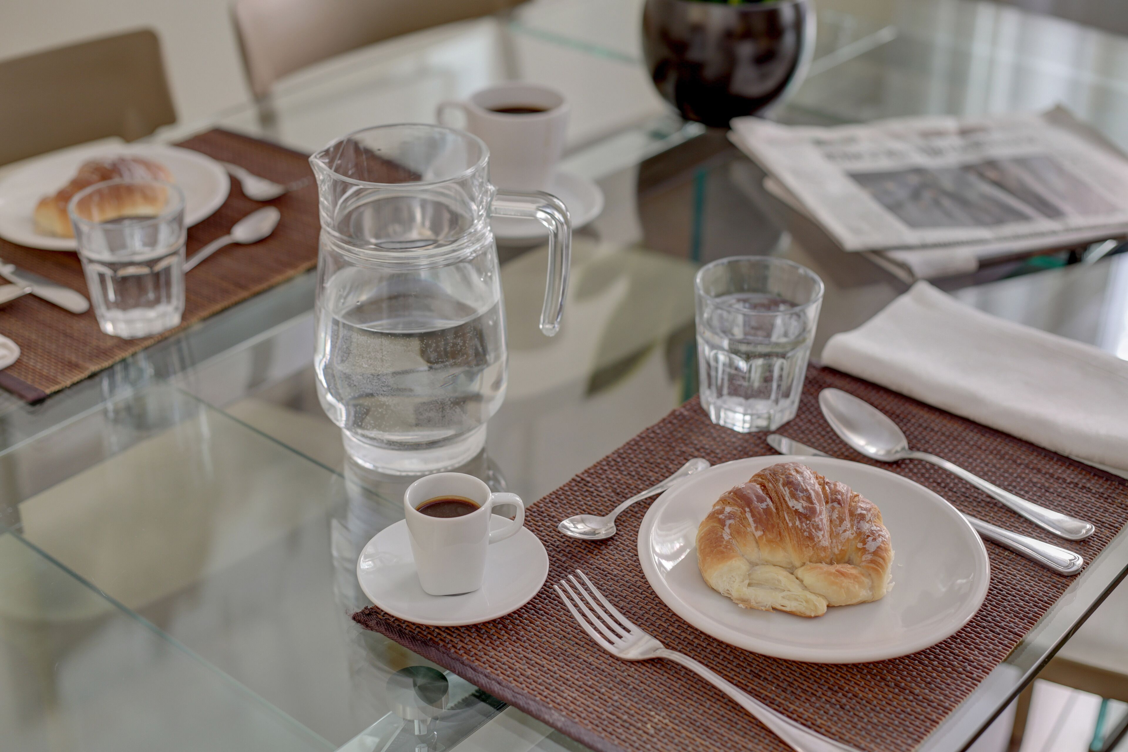 Un esmorzar amb un croissant, cafè, gots d'aigua i una testosseta sobre una taula de vidre.