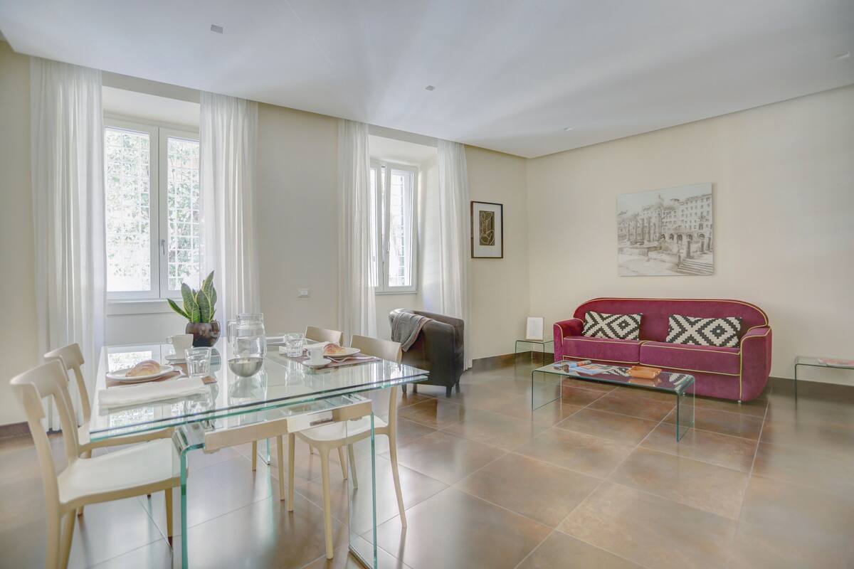 A modern living room with a glass dining table, a sofa, and a large window with curtains.