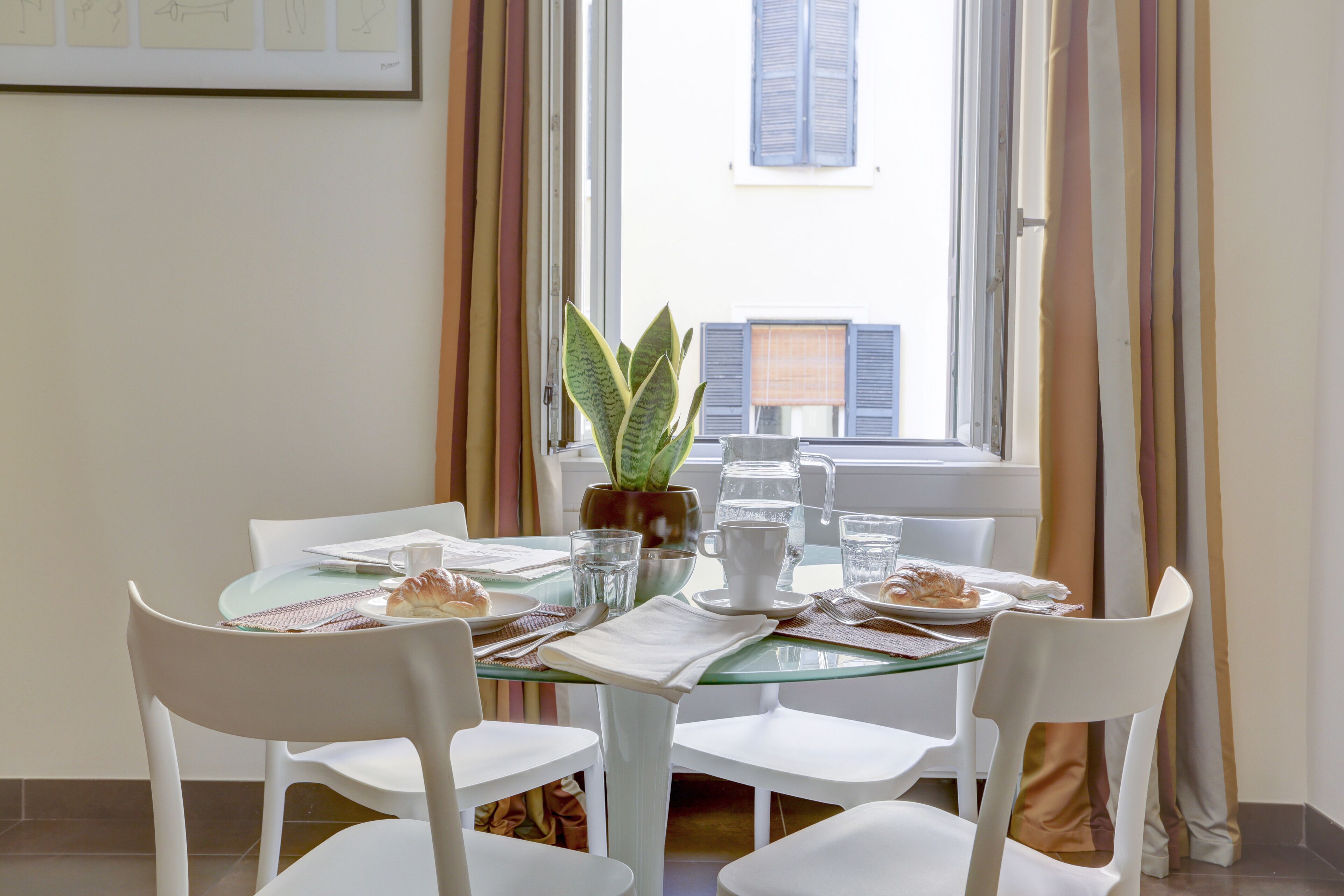 A dining area with a glass table, white chairs, a potted plant, and breakfast items.