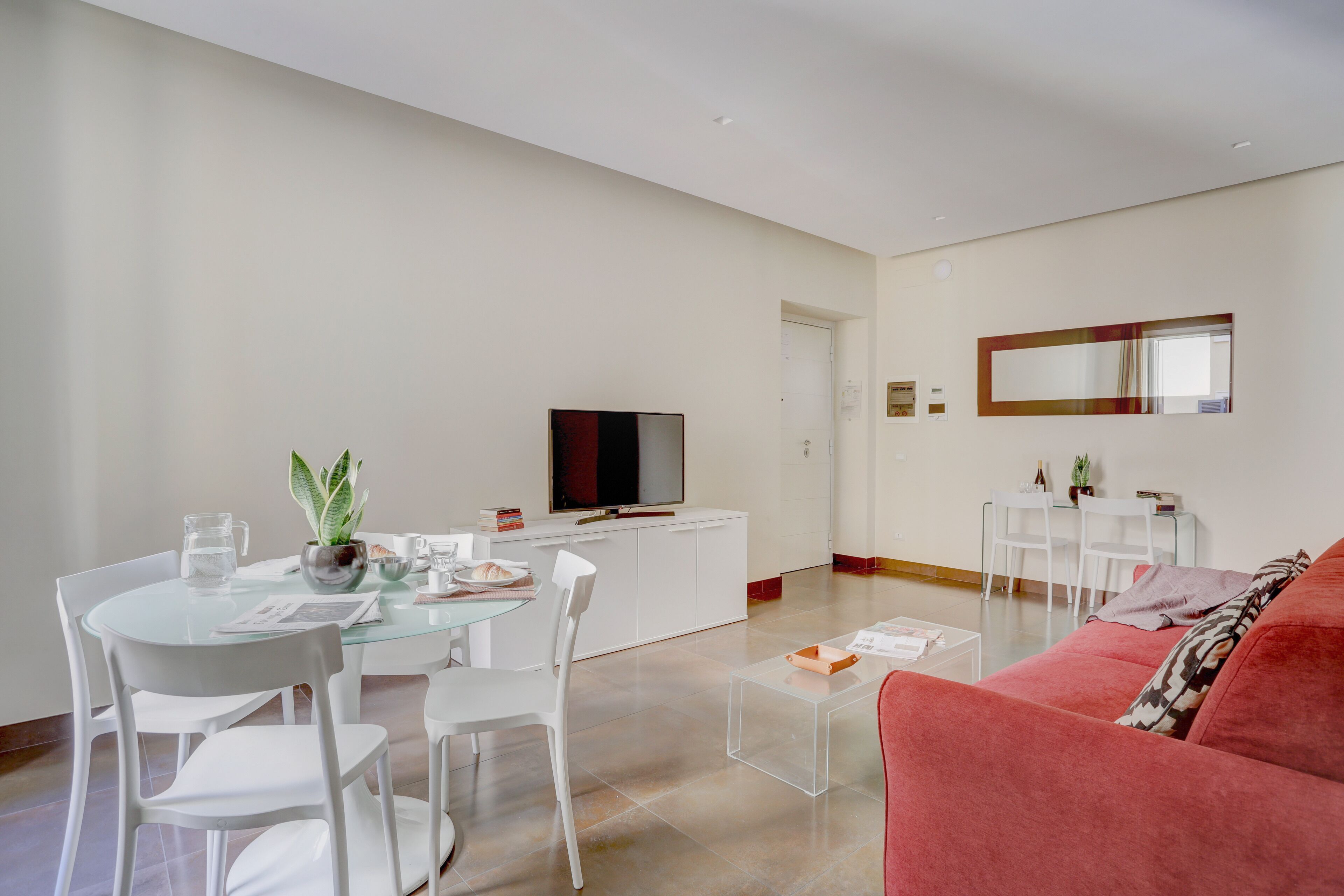 Ein modernes Wohnzimmer mit einem Glastisch, weißen Stühlen, einem roten Sofa und einem Flachbildfernseher auf einer weißen Kommode.