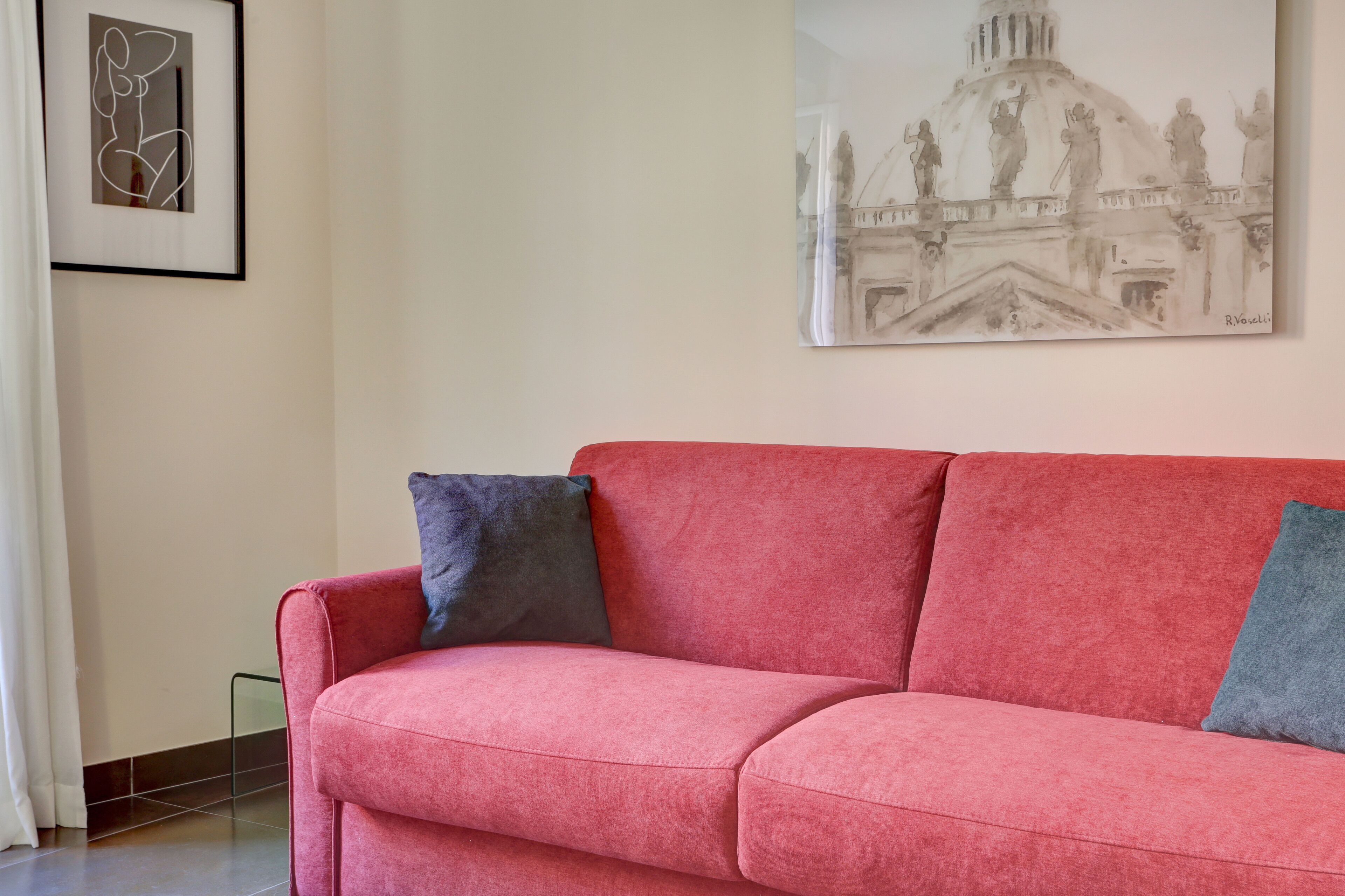 A red sofa with two pillows, a framed abstract artwork, and a classical building painting on the wall.