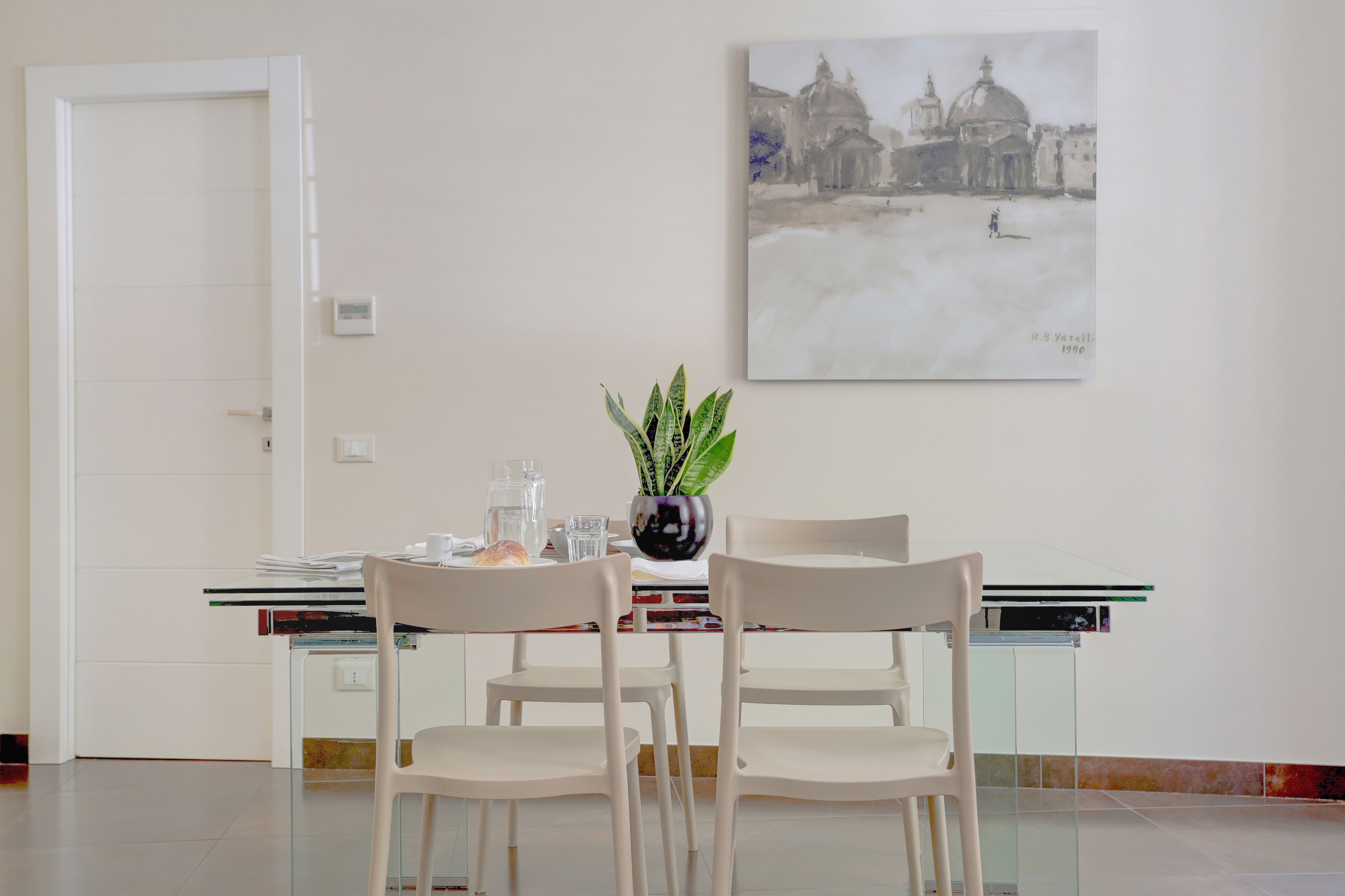 A dining area with a table set for two, a potted plant, and a painting on the wall.