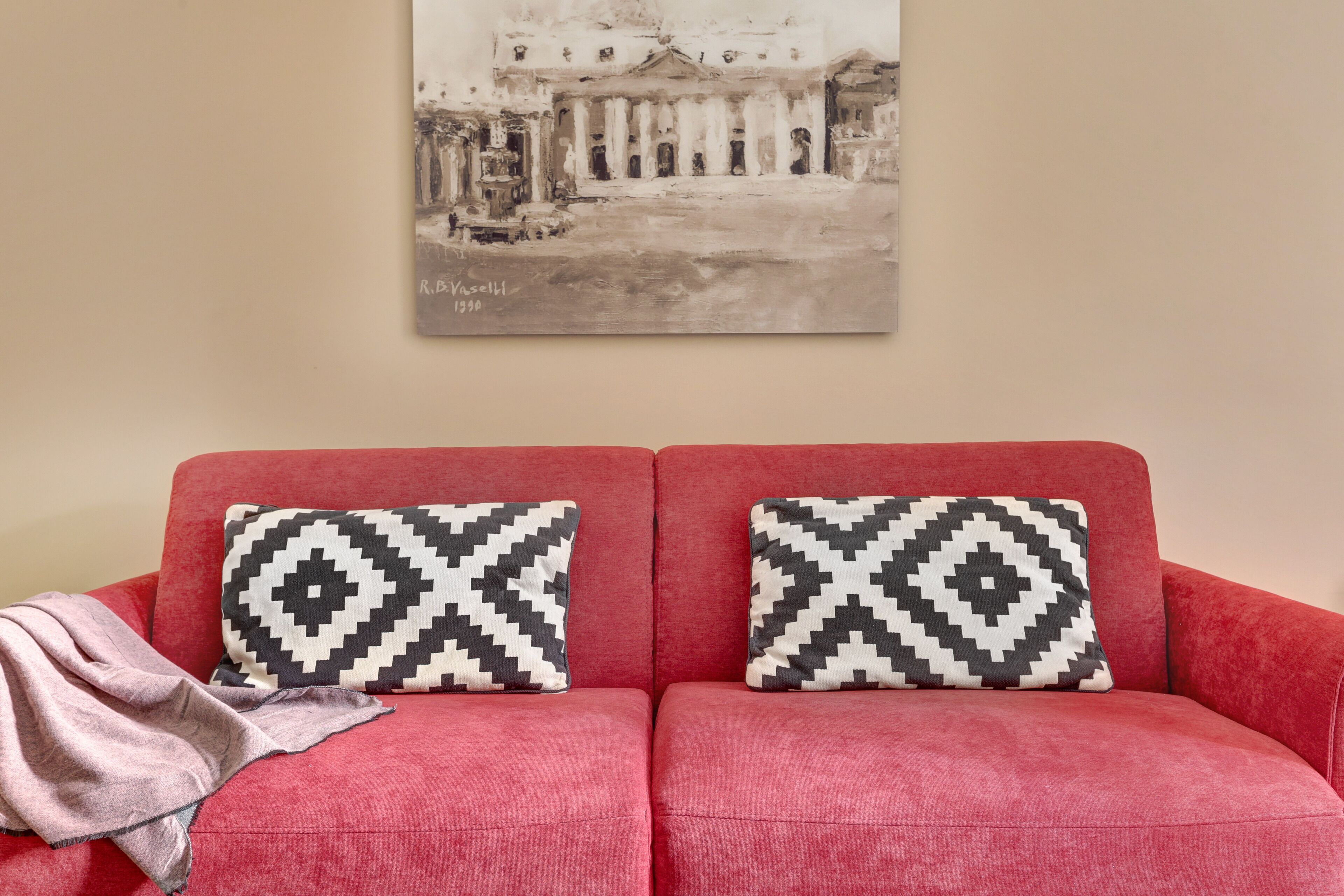 A red sofa with two patterned pillows and a blanket, a framed artwork above it, and a plain wall.