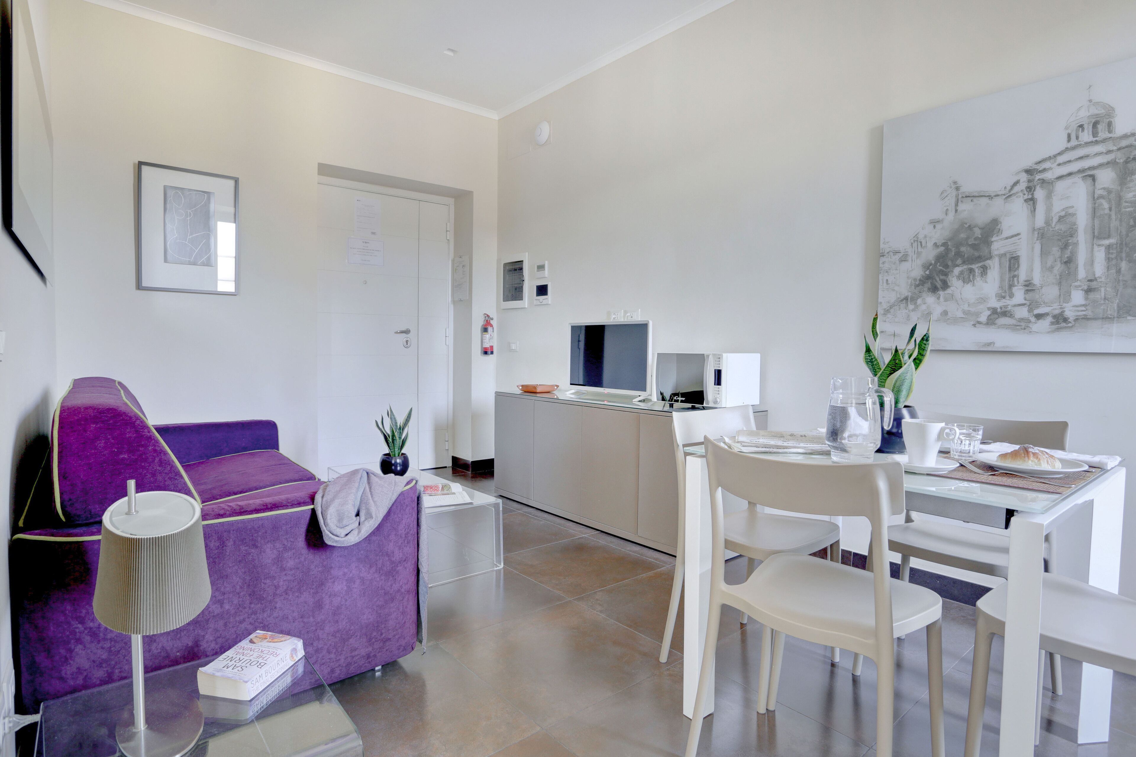 A modern living room with a purple sofa, a small table with a lamp, and a dining area with chairs and a table set for two.