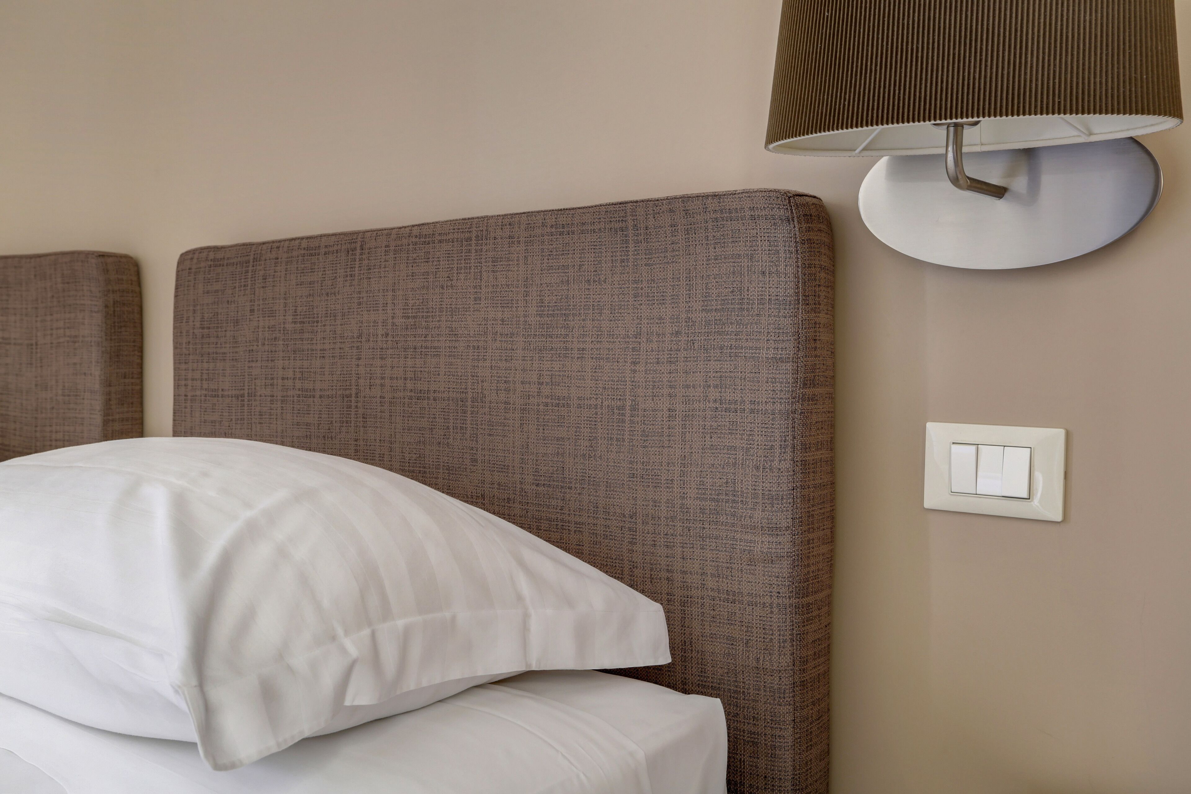 Una cama bien tendida con ropa blanca, un aplique en la pared y un interruptor de luz en una pared color beige.