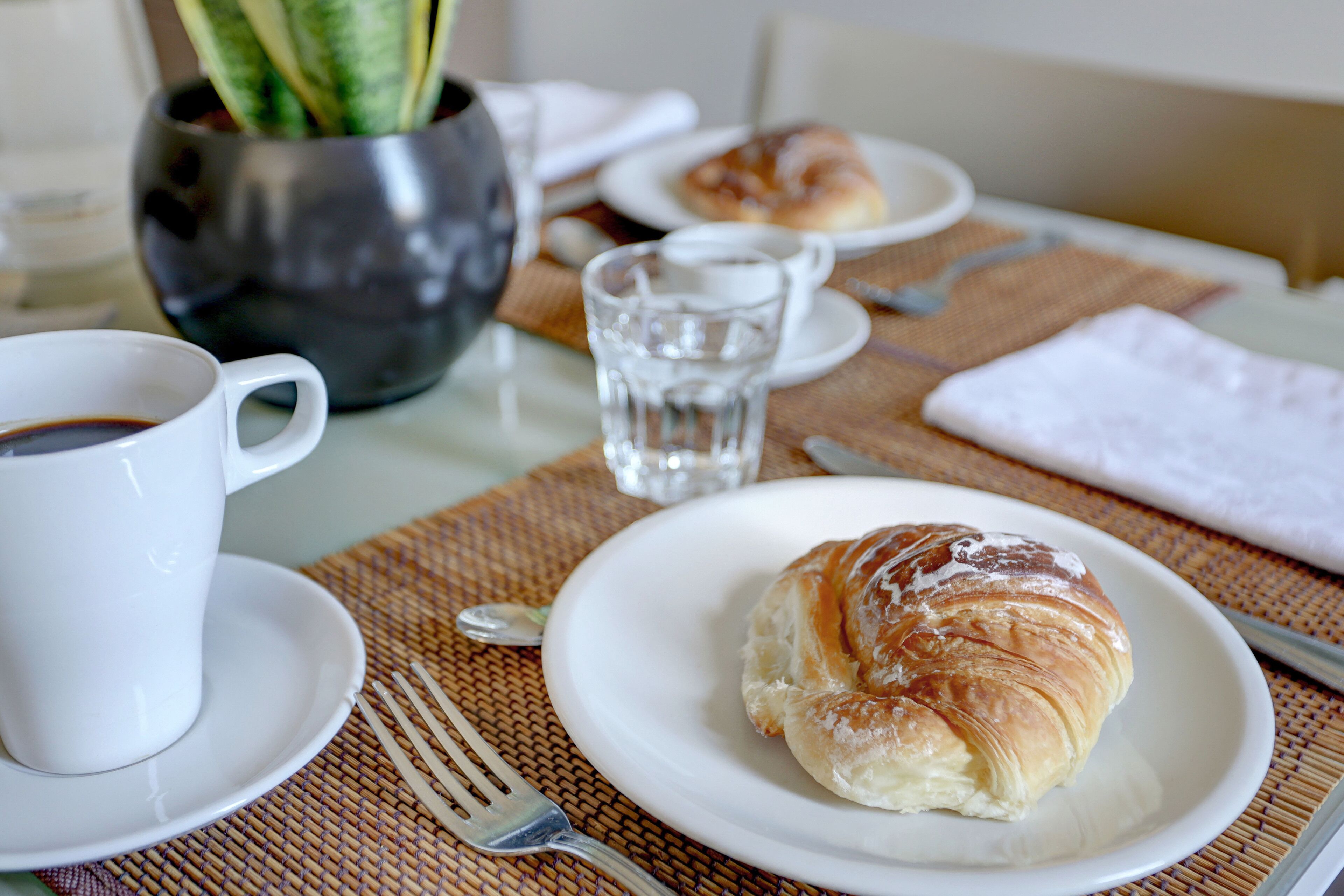 A breakfast setting with a croissant on a plate, a cup of coffee, and a glass of water on a table.