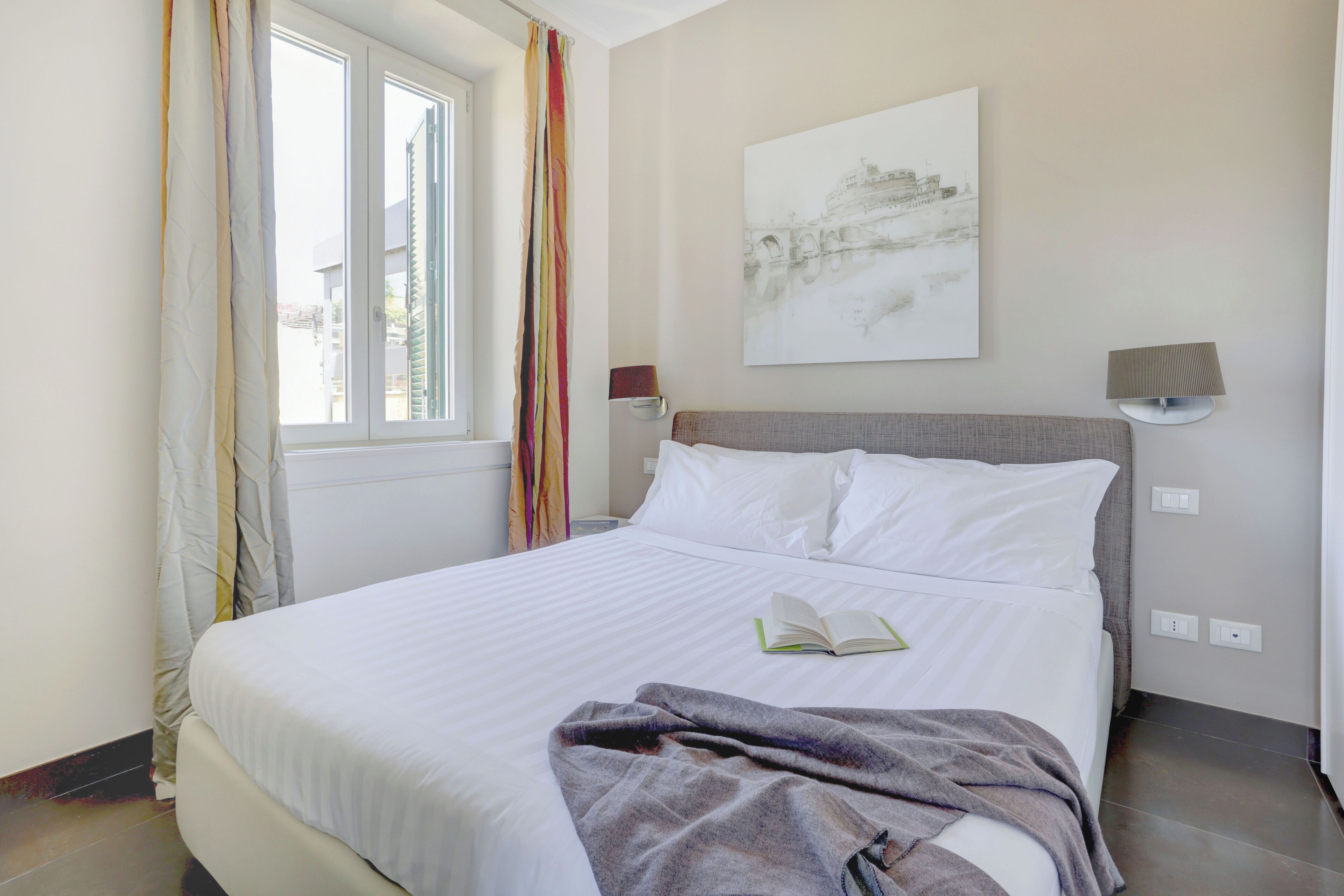 A neatly made bed with white linens, a book on it, and a colorful curtain partially drawn.