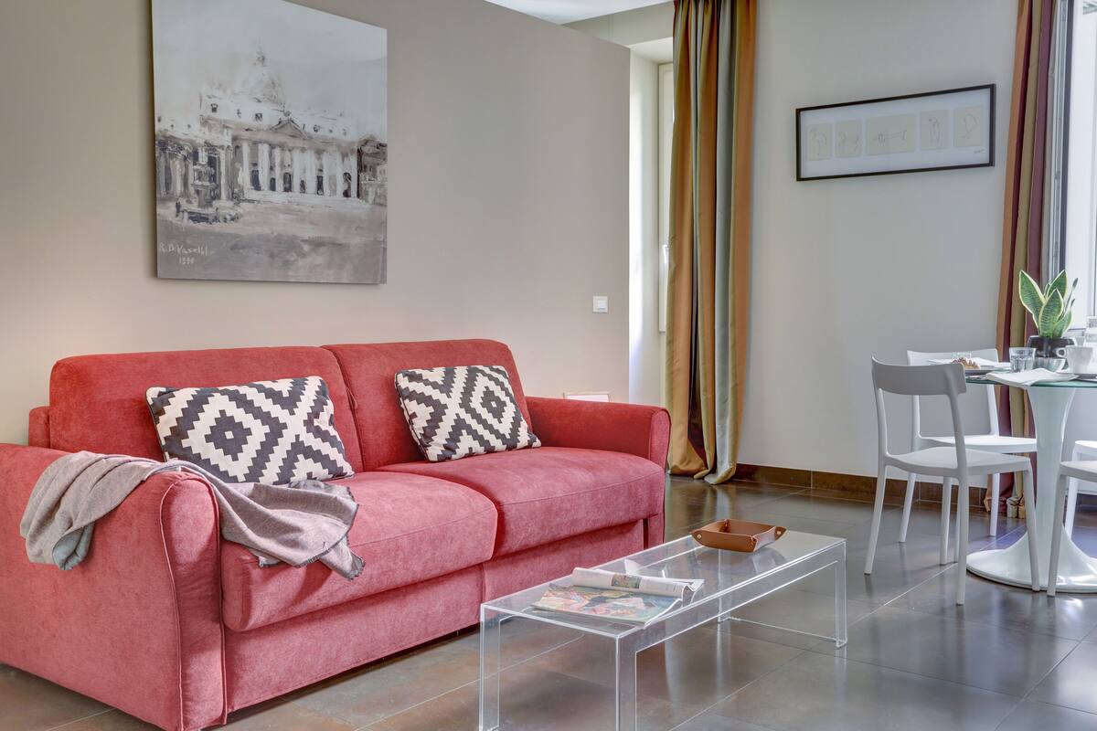 A living room with a red sofa, a clear coffee table, and a dining area with white chairs and a table.