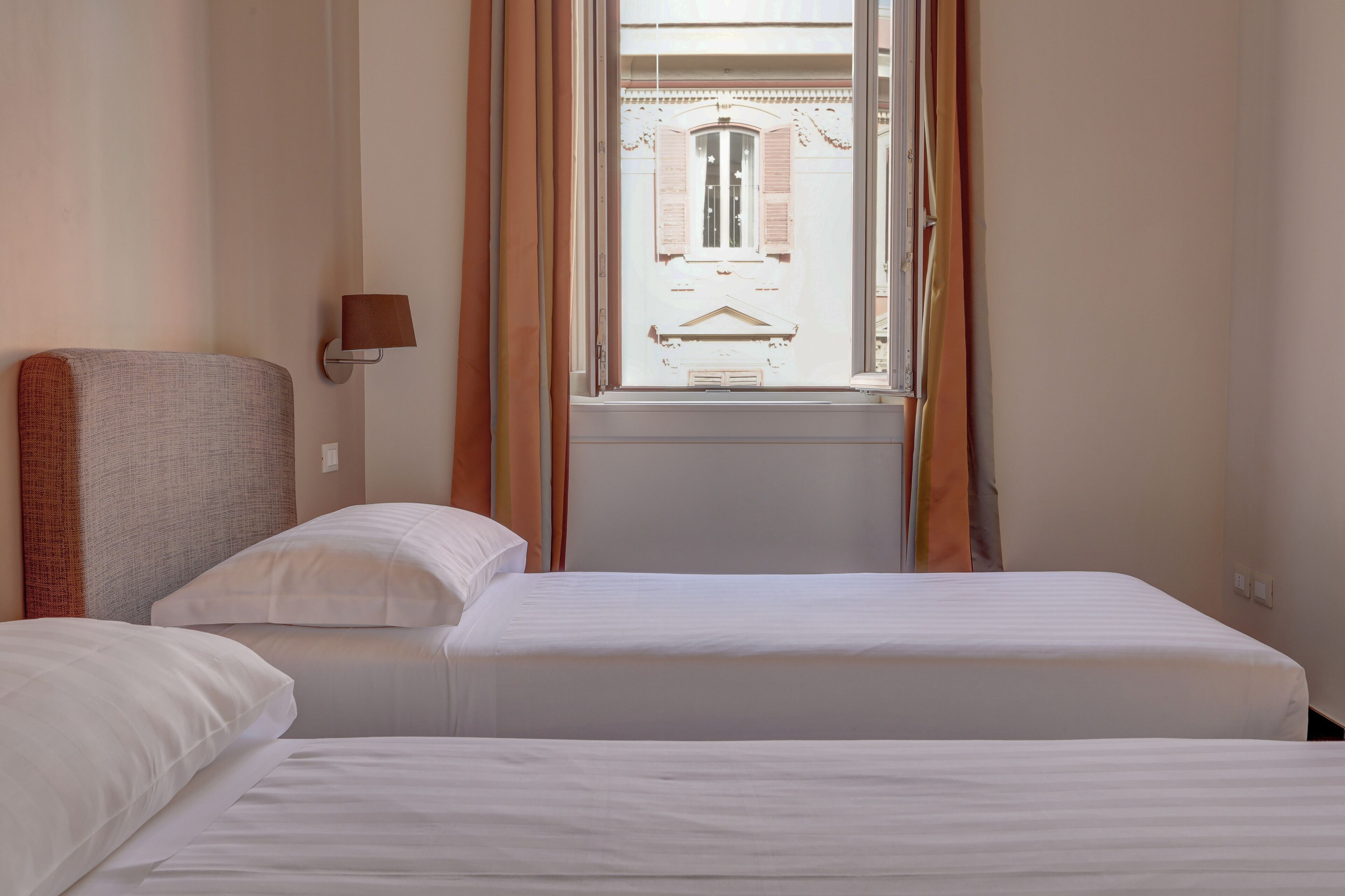 Une chambre d’hôtel avec deux lits, une tête de lit, une fenêtre avec des rideaux et une lampe fixée au mur.