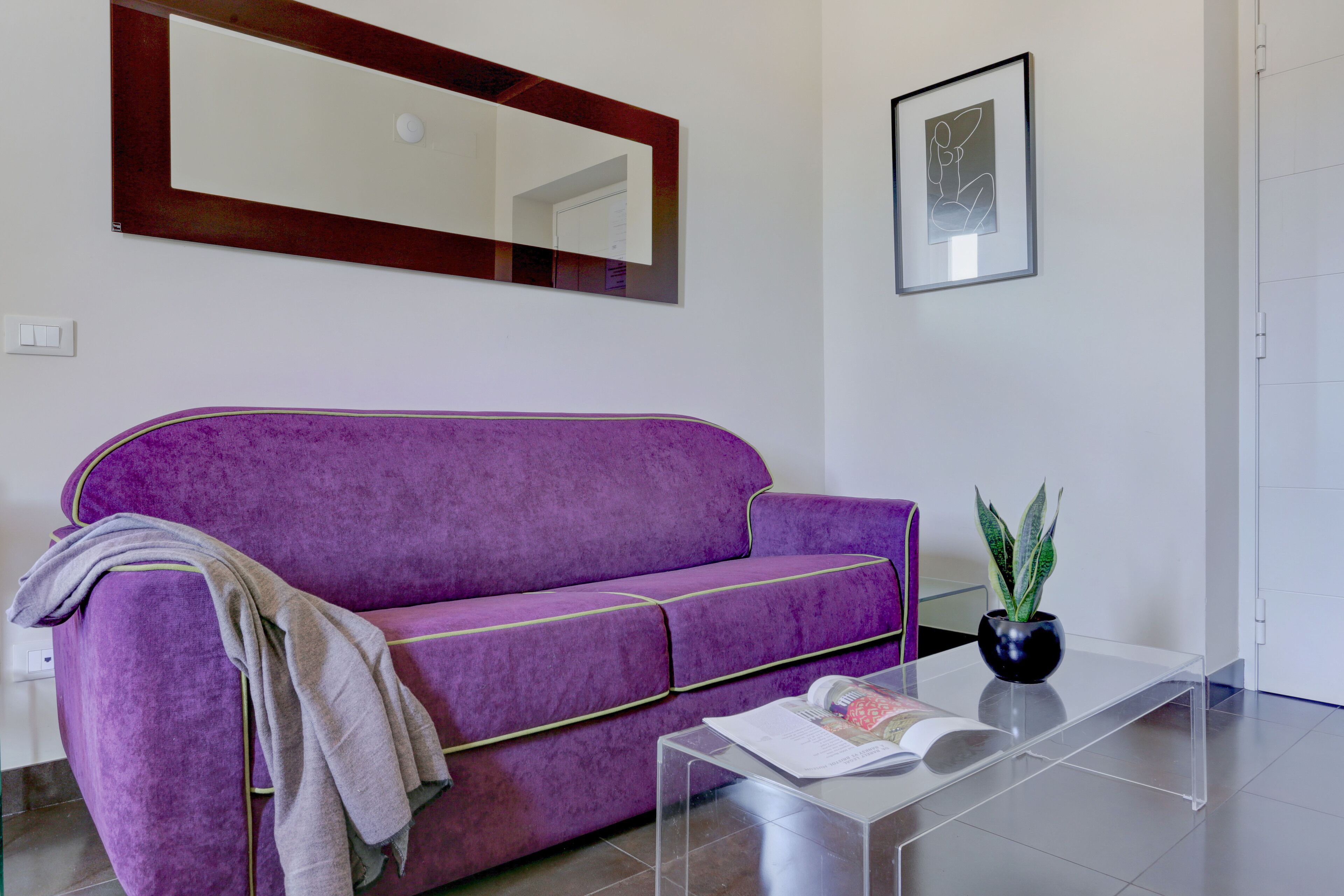 A purple sofa with a throw blanket, a clear glass coffee table with a magazine, a potted plant, and a framed abstract artwork on the wall.
