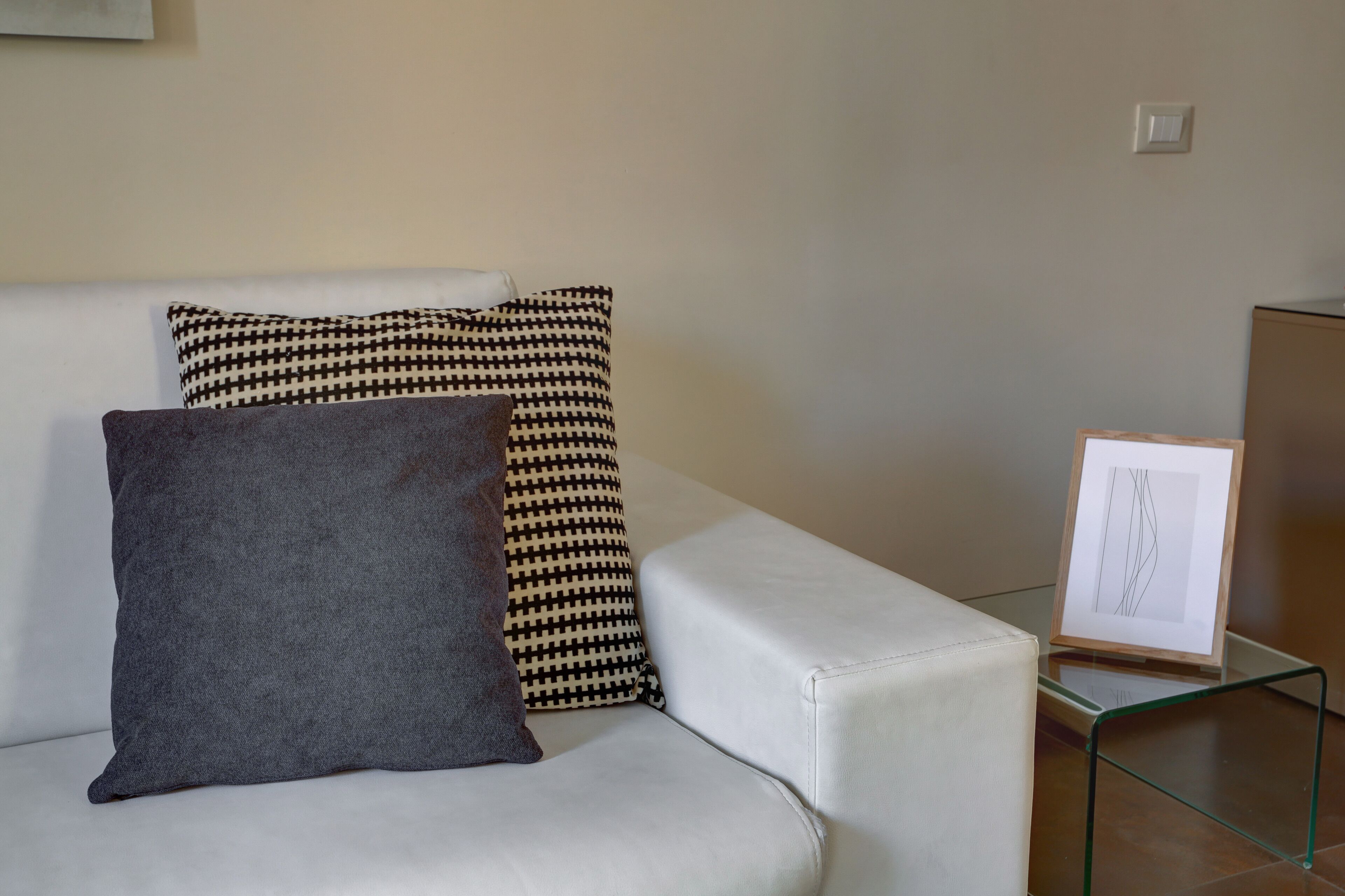 A modern living room with a white sofa, patterned cushions, and a framed artwork on a glass table.