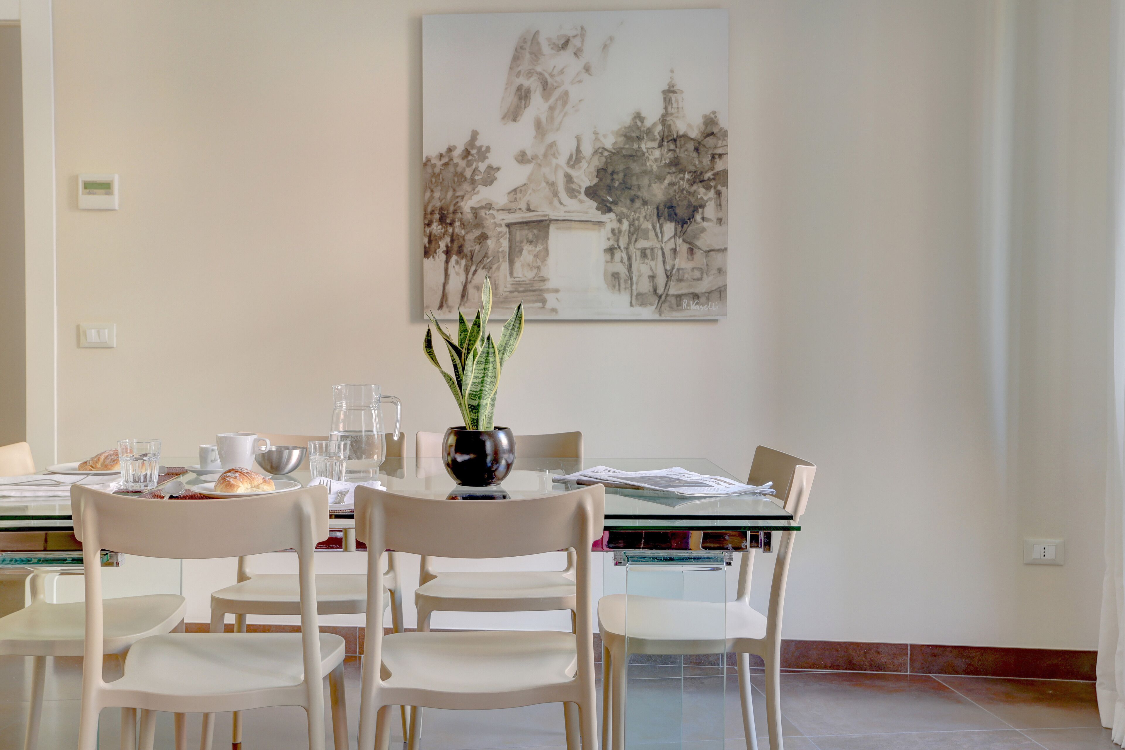 A dining area with a table set for two, a potted plant, and a painting on the wall.
