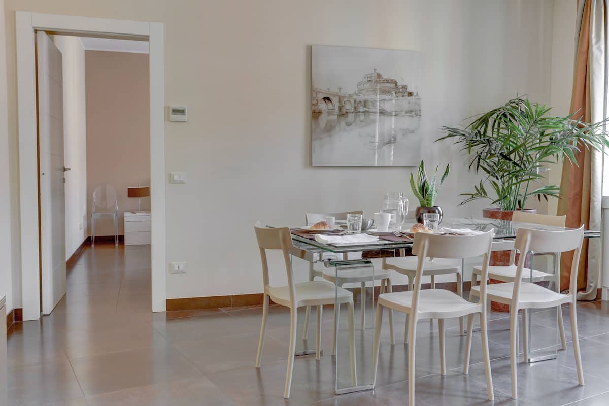 A dining area with a glass table, white chairs, a potted plant, and a wall-mounted artwork.