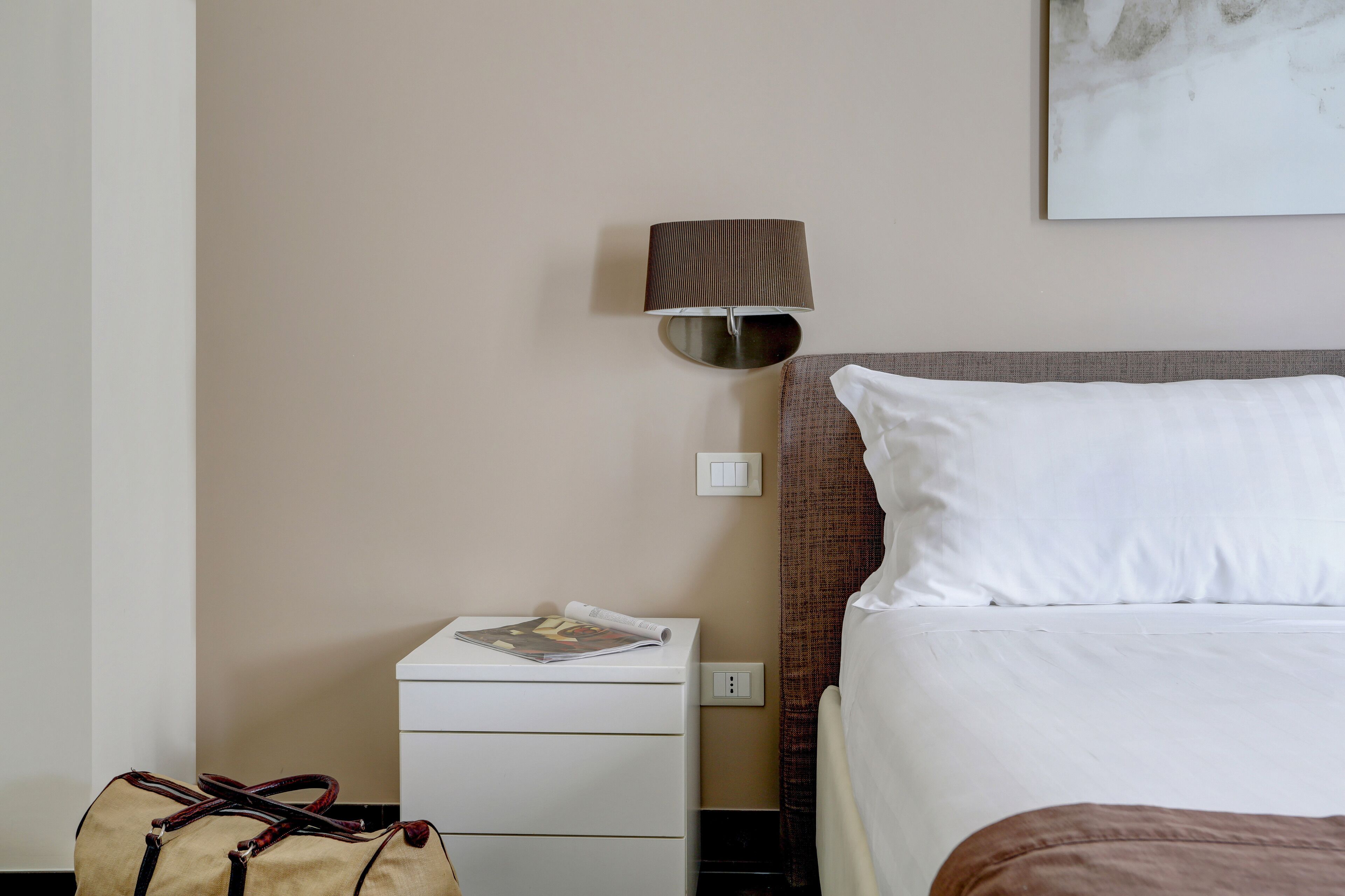 A hotel room with a bed, a nightstand, a lamp, a magazine, and a bag.