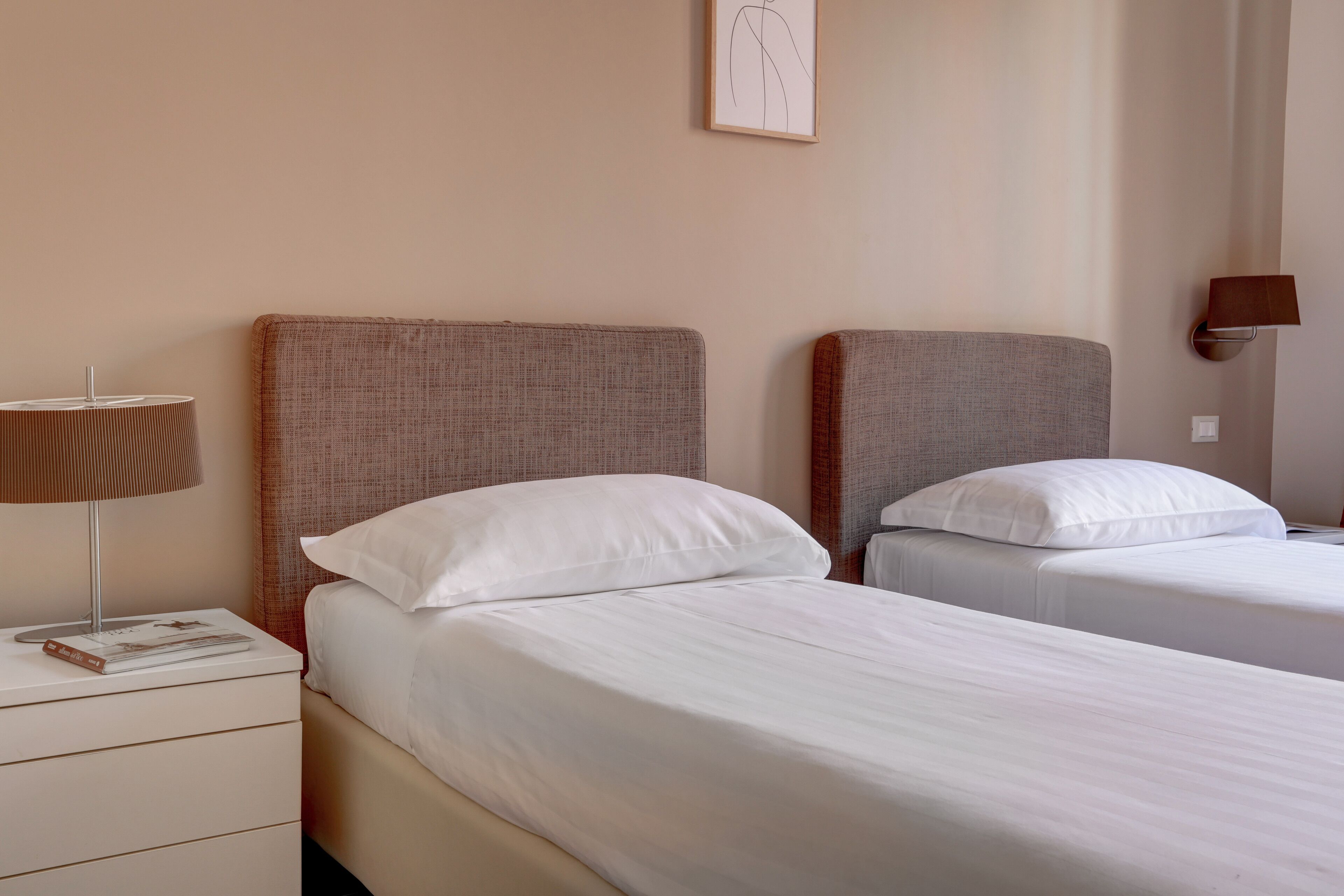 A hotel room with two beds, a bedside table with a lamp, and a framed picture on the wall.