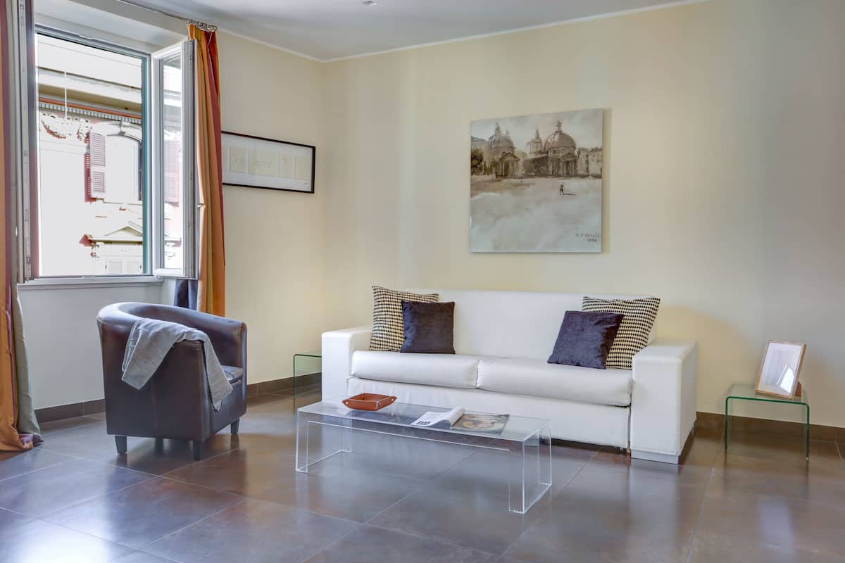 A living room with a white sofa, a brown armchair, a glass coffee table, and a painting on the wall.