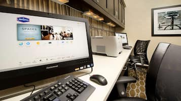 Un espace de travail aménagé dans une chambre d’hôtel, avec un grand écran affichant une publicité pour Hampton Spaces, un ordinateur Lenovo, un clavier, une souris et des chaises de bureau.