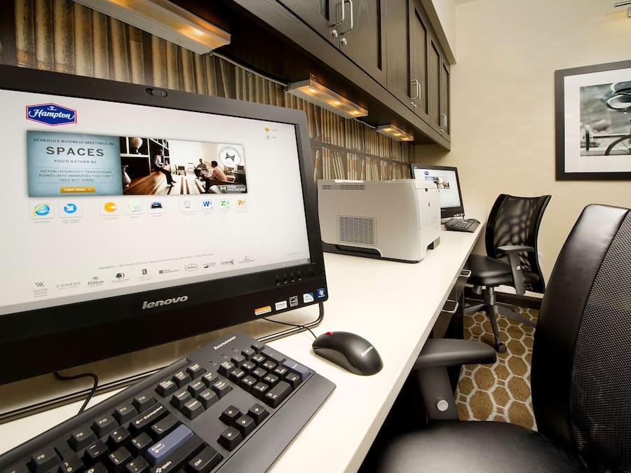 Un espace de travail aménagé dans une chambre d’hôtel, avec un grand écran affichant une publicité pour Hampton Spaces, un ordinateur Lenovo, un clavier, une souris et des chaises de bureau.