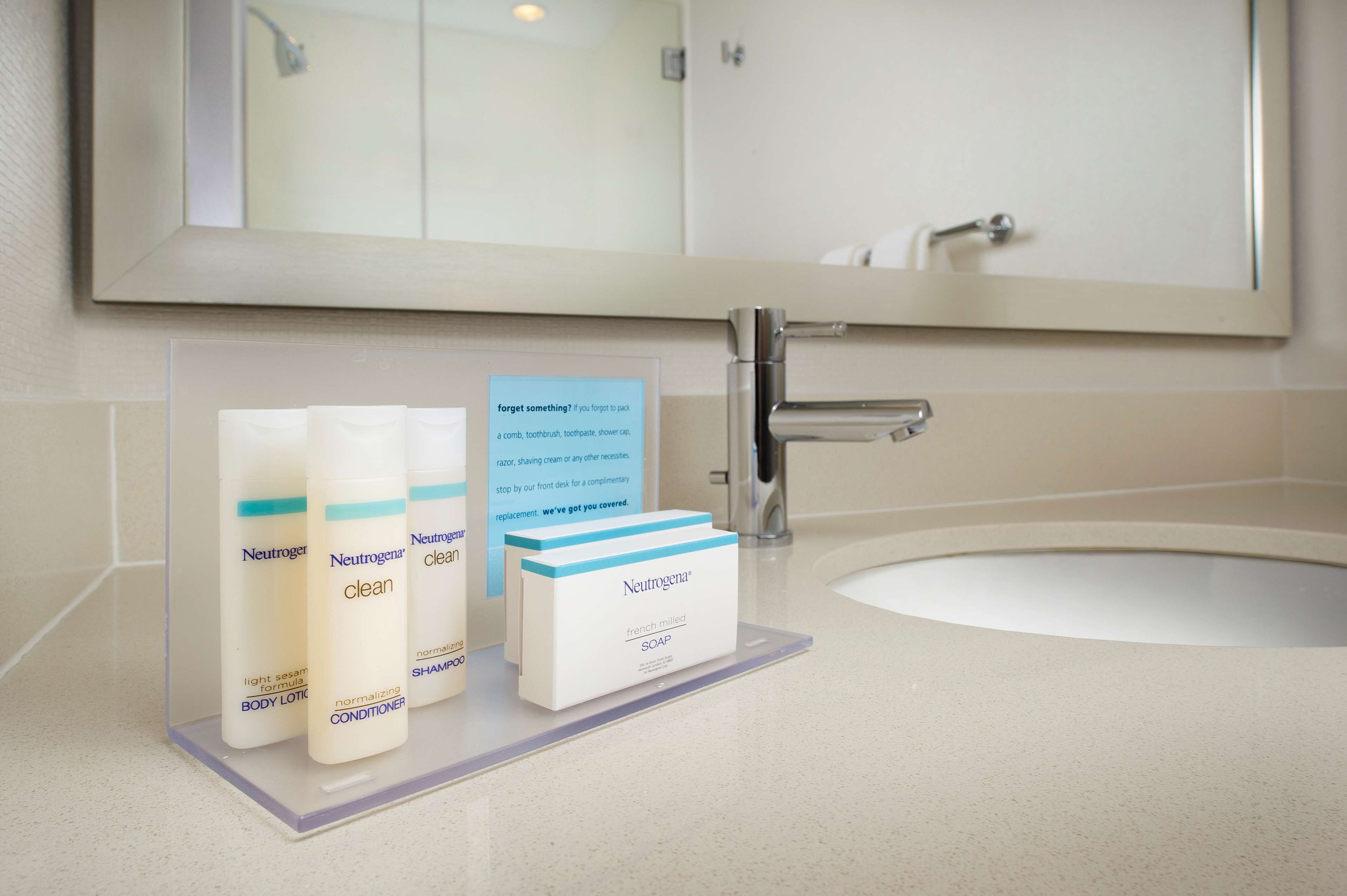 Un baño con lavamanos, espejo y artículos de aseo, incluyendo loción corporal, acondicionador y jabón Neutrogena.